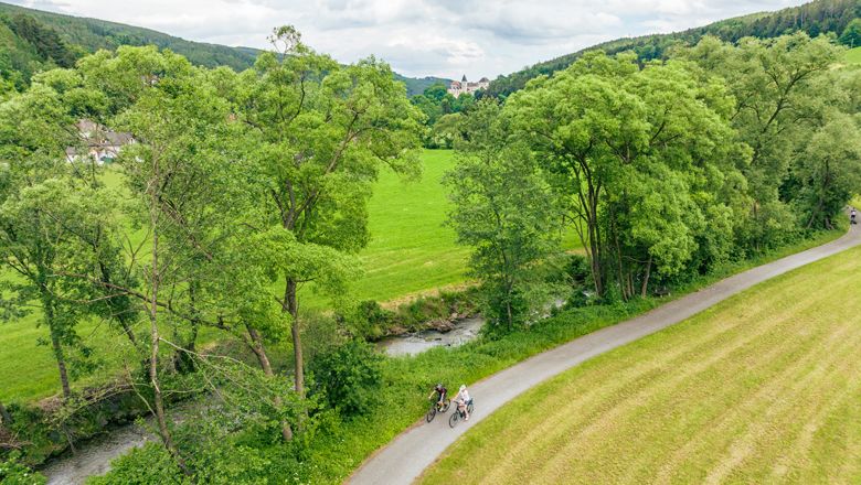 Dva cyklisté na stezce podél řeky obklopené zelenými stromy a loukami.