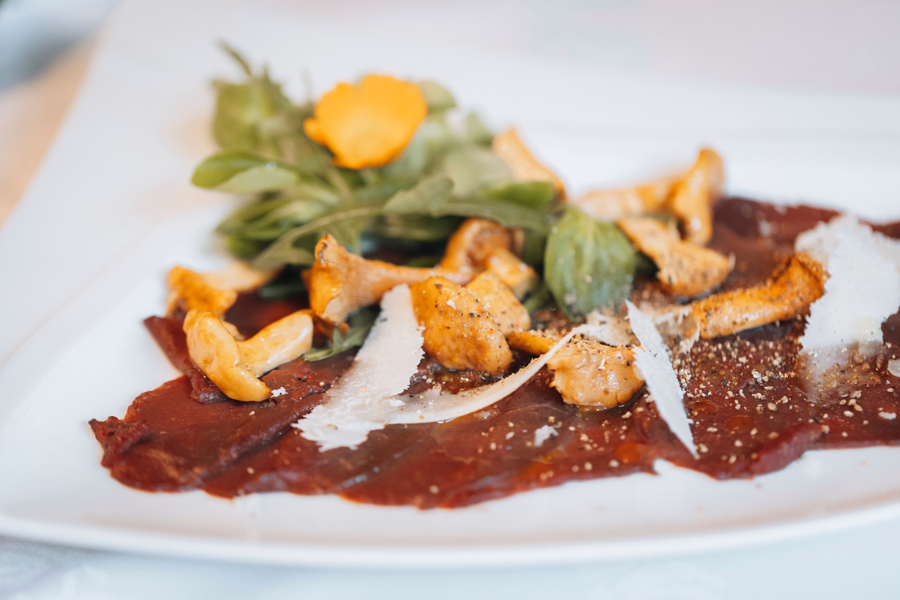 Carpaccio z daňka s nakládanými liškami a rukolou na bílém talíři.