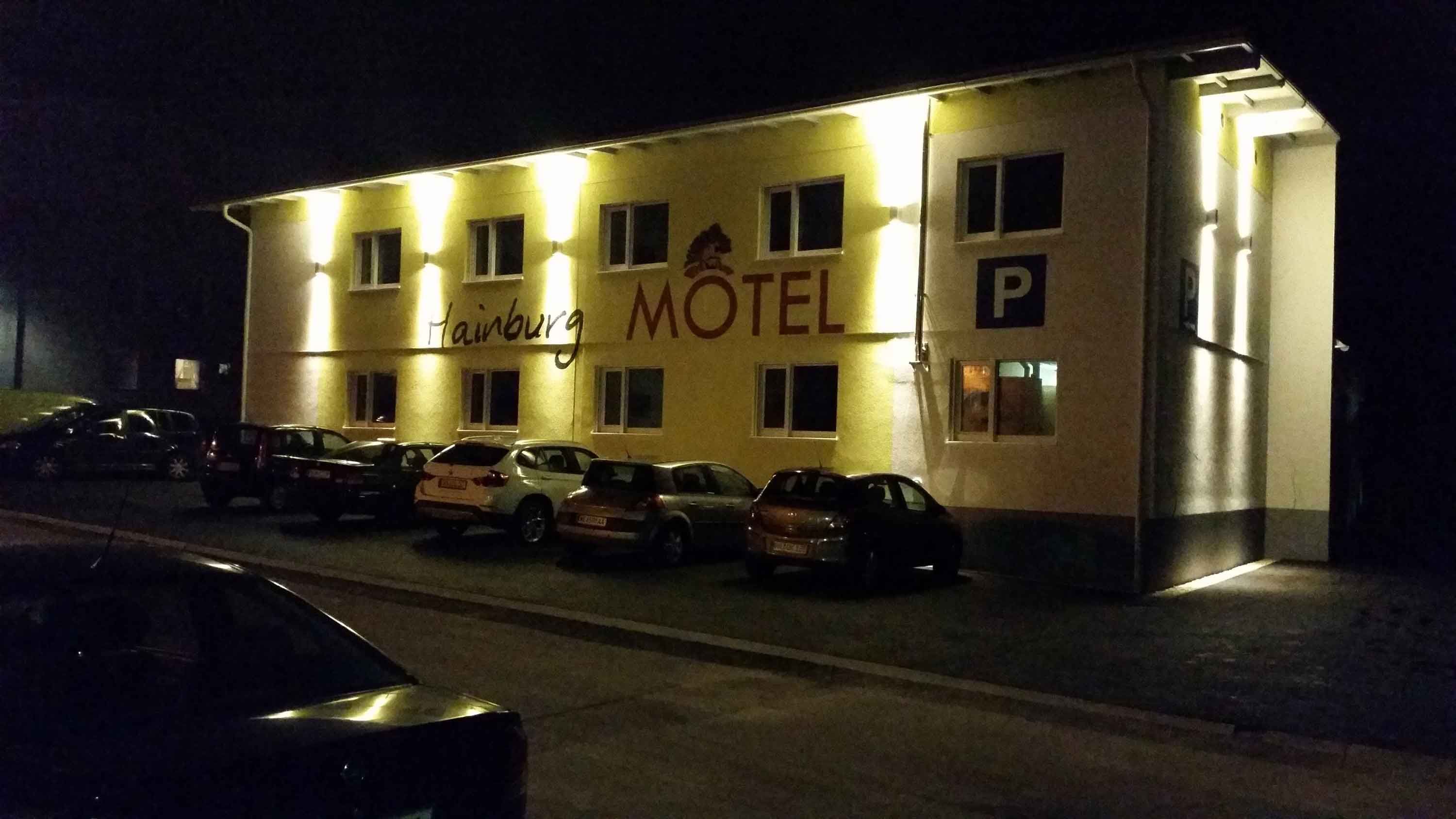 Noční záběr osvětleného motelu s nápisem "Hainburg Motel" a zaparkovanými auty před ním.