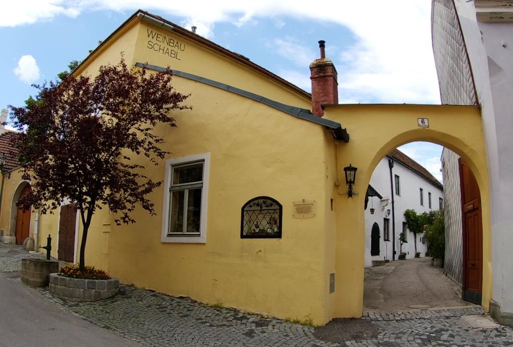 Žlutá budova s nápisem "Weinbau Schabl", vedle ní oblouk a strom.
