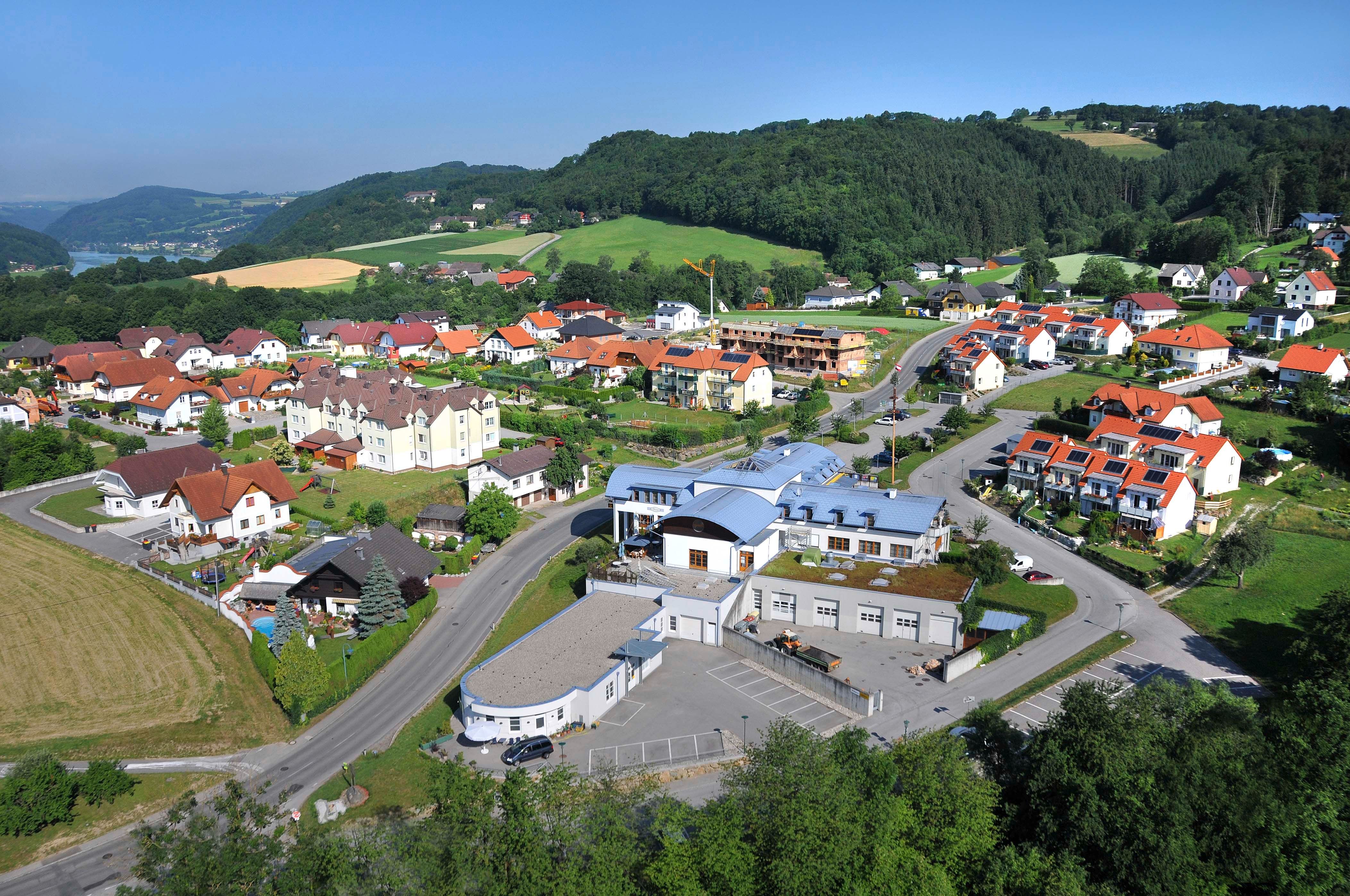 Letecký pohled na vesnici s komunitním centrem a okolními domy v zelené kopcovité krajině.
