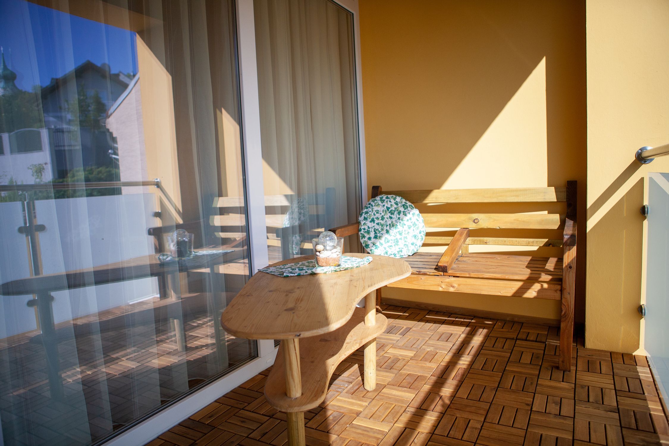 Balkon s dřevěnou podlahou, dřevěným stolem a dřevěnou lavicí s kulatým polštářem. V pozadí je vidět skleněné zábradlí a budova.