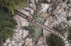 Zwei Wanderinnen gehen über eine schmale Holzbrücke, die über einen klaren, von Felsen umgebenen Fluss in einer Schlucht führt