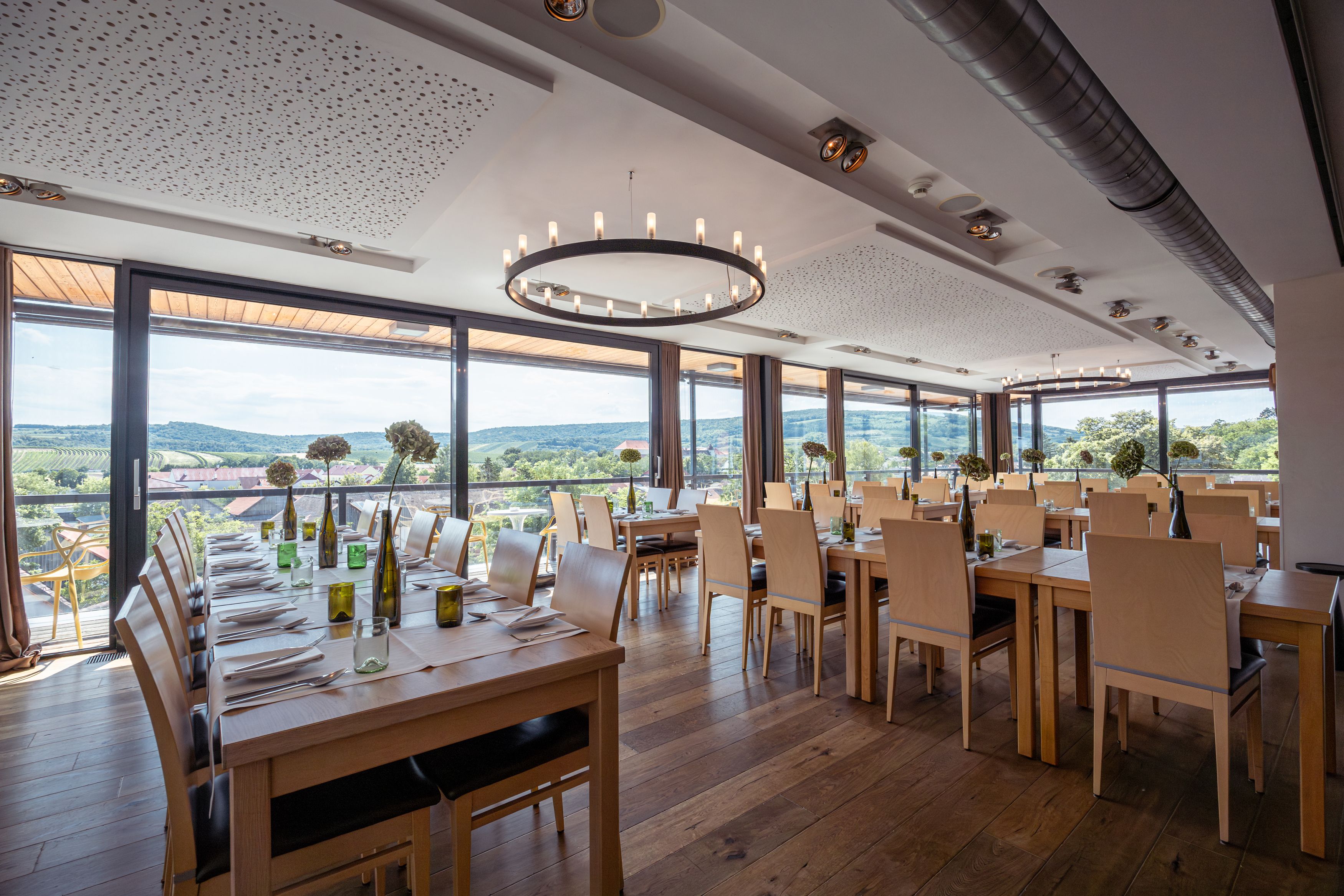 Pohled do interiéru moderní restaurace s velkými okny a výhledem na vinice.