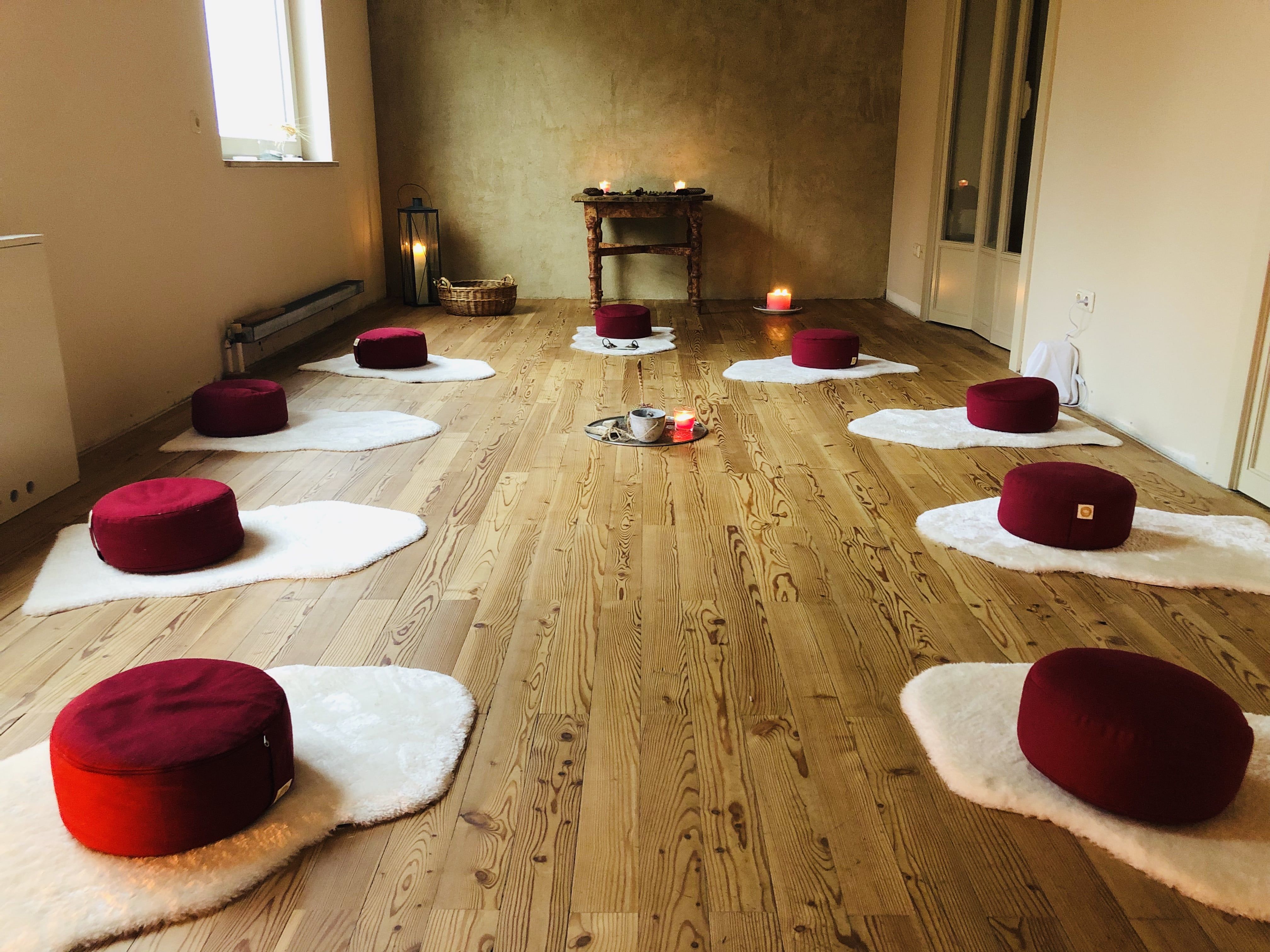 Meditační místnost s červenými polštáři na bílých rohožích, svíčkami a dřevěnou podlahou.