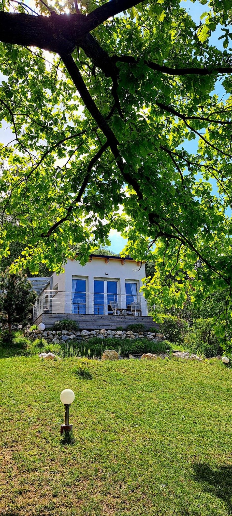 Moderní chata s velkým oknem, obklopená zelení a stromy.