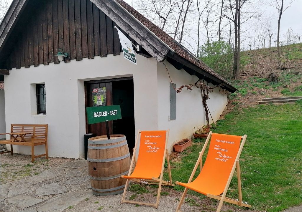 Malá bílá budova s dřevěnou střechou, dvěma oranžovými lehátky a dřevěným sudem před ní, obklopená stromy a trávou.
