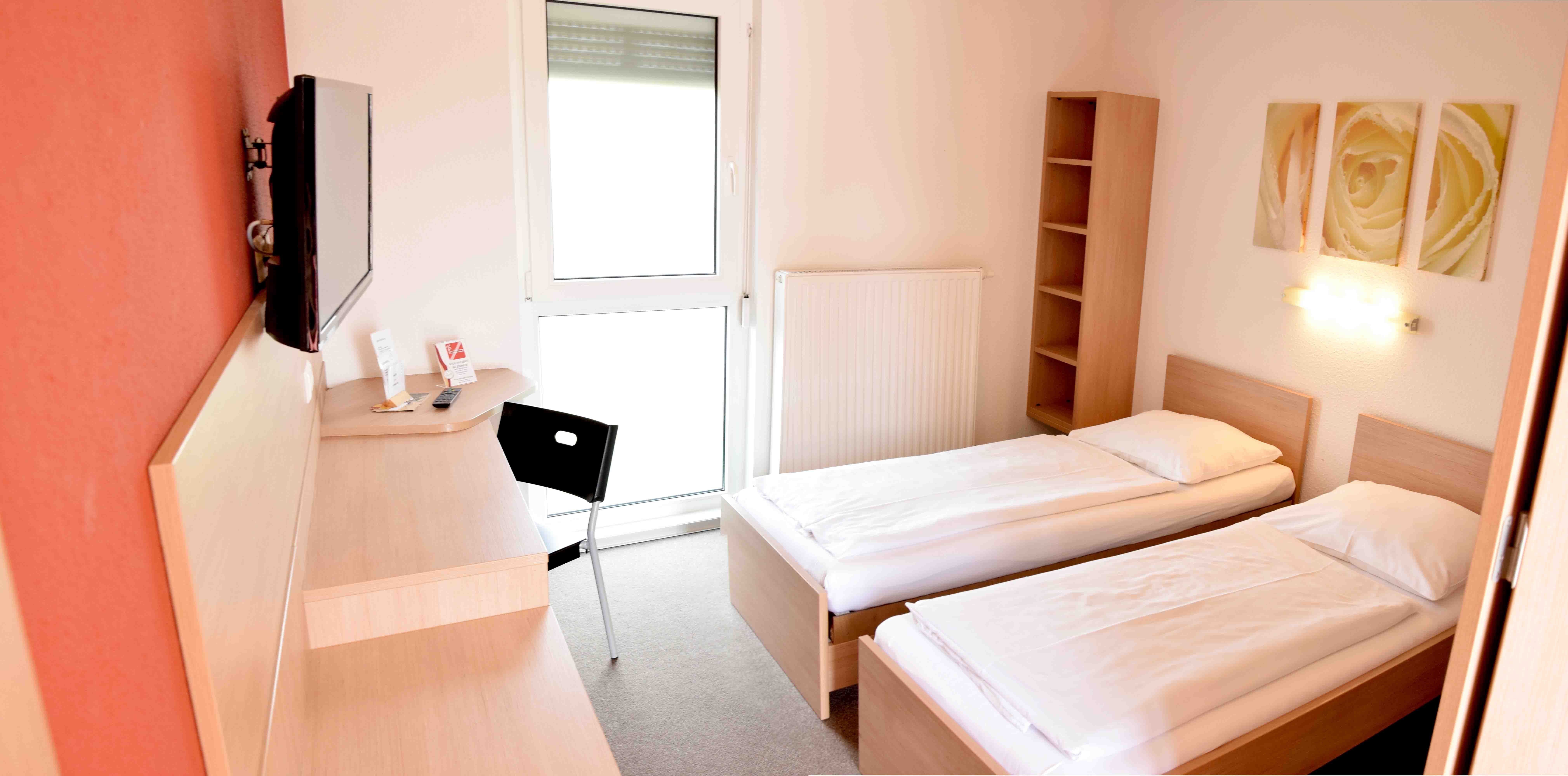 Jednoduchý motelový pokoj se dvěma samostatnými postelemi, stolem, židlí a televizí na stěně.