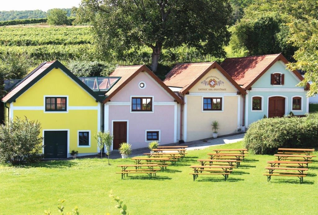 Tři barevné budovy vinařství Brunthaler s pivními lavicemi v popředí a vinicemi v pozadí.