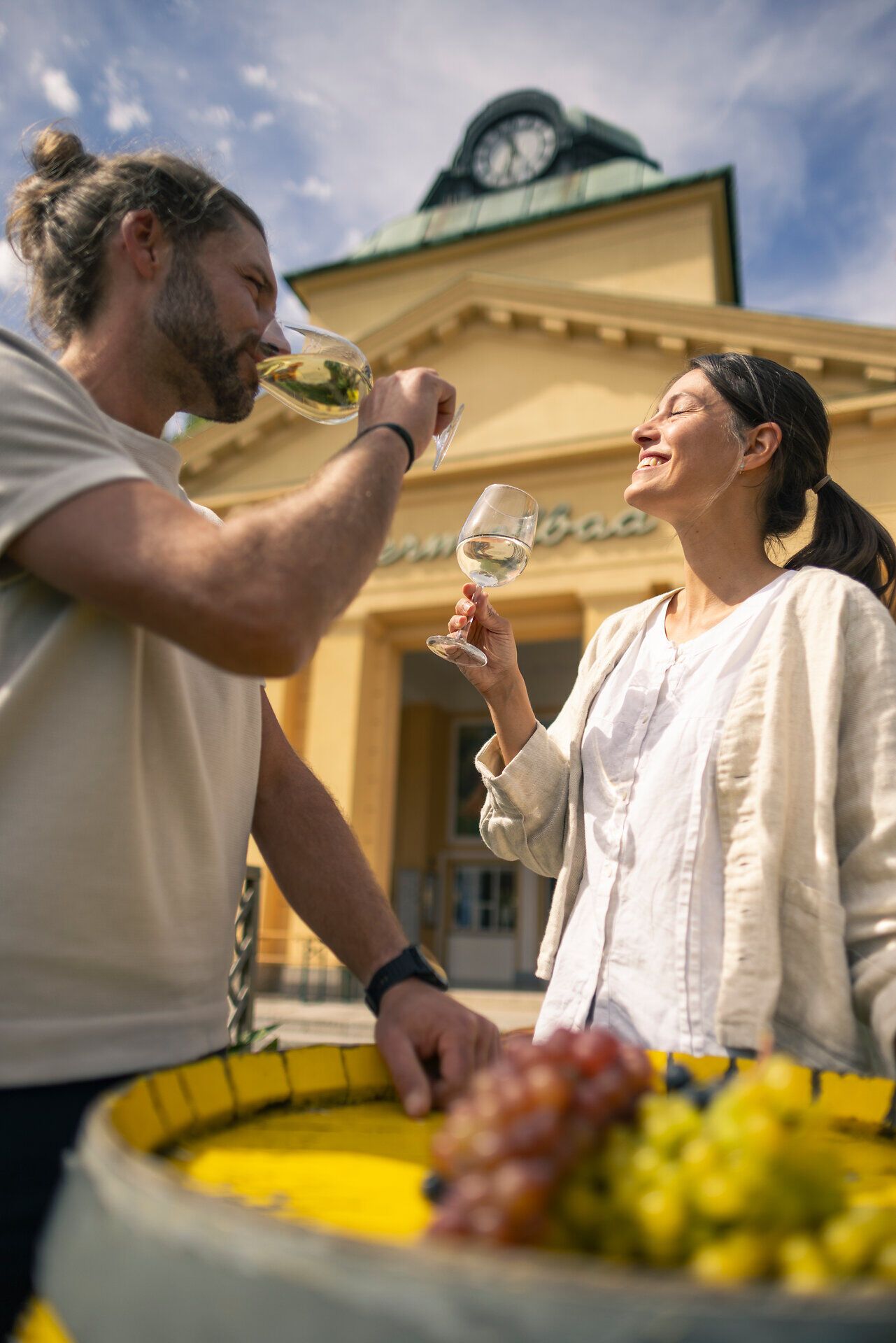 Vor dem Thermalbad Bad Vöslau stehen ein Mann und eine Frau vor einem mit Trauben verzierten Fass. Beide halten ein Glas Wein in der Hand, während die Frau die Sonne genießt. Die Kulisse im Wienerwald strahlt Ruhe und Naturverbundenheit aus.