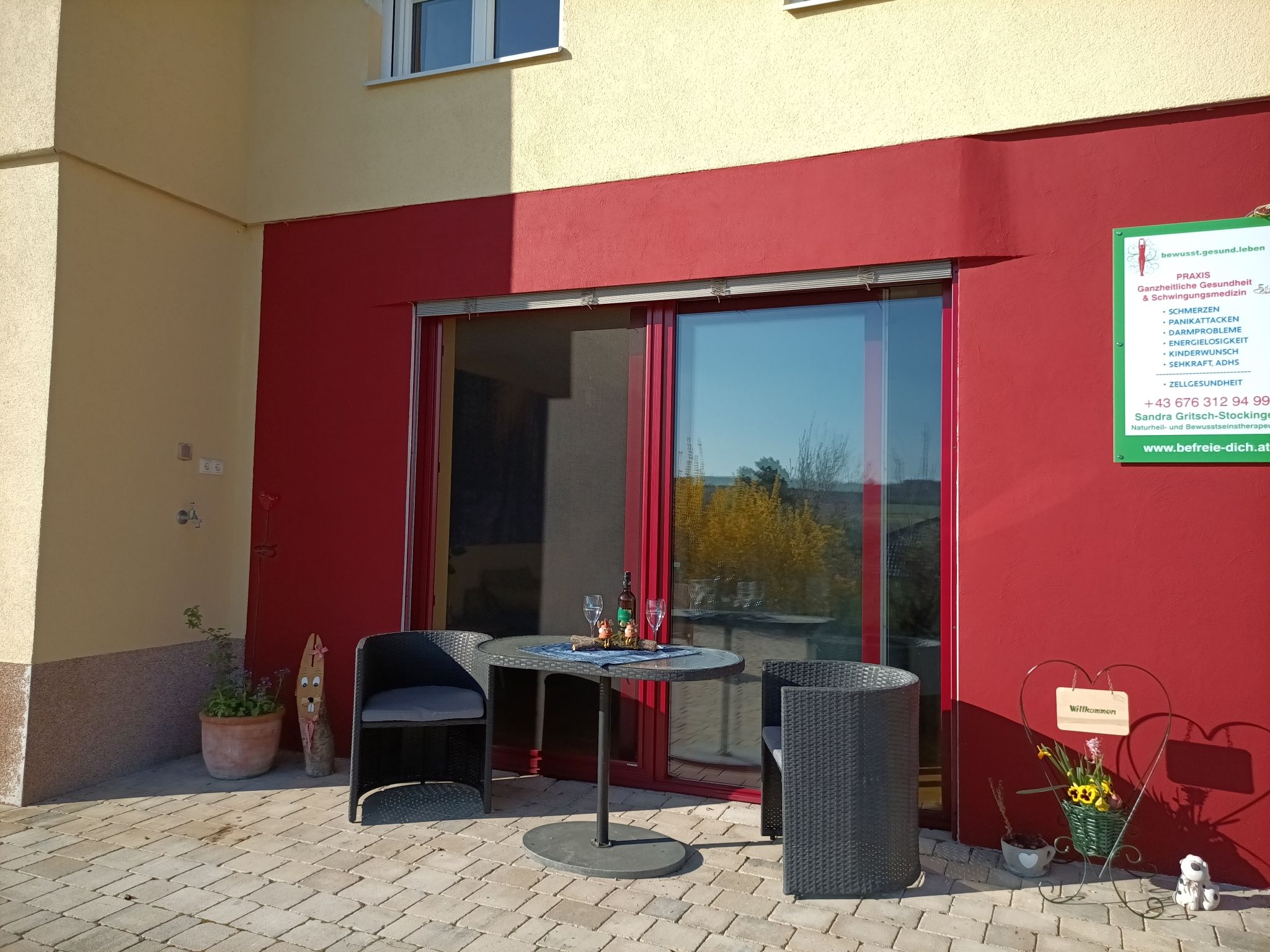 Terasa se dvěma židlemi a stolem před červenou zdí, vedle cedule s informacemi.