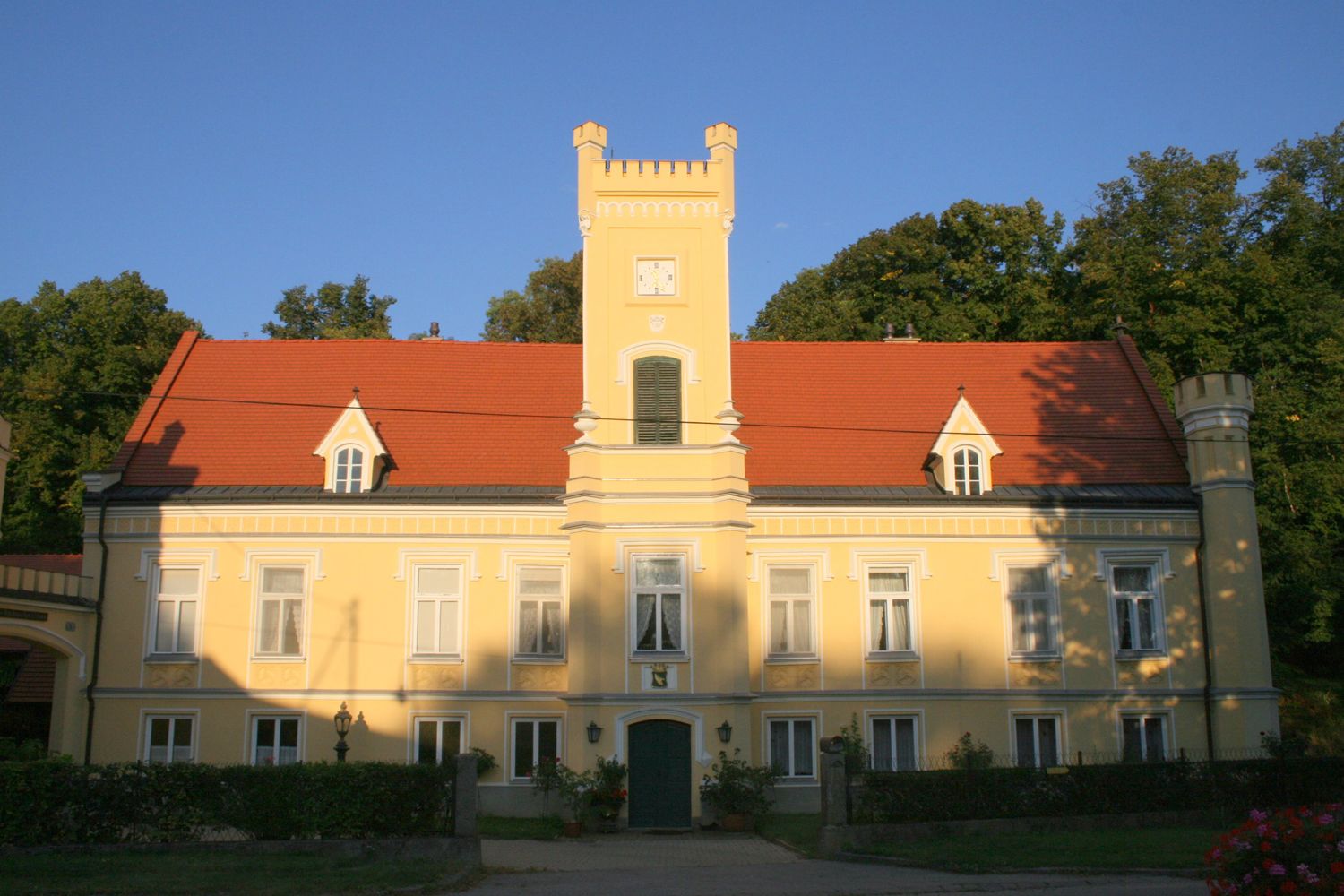 Žlutý hrad s červenou střechou a věží, obklopený stromy.