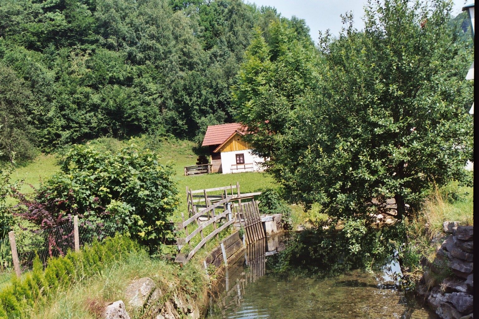 V zelené krajině vedle potoka a stromů stojí malá chata s červenou střechou.
