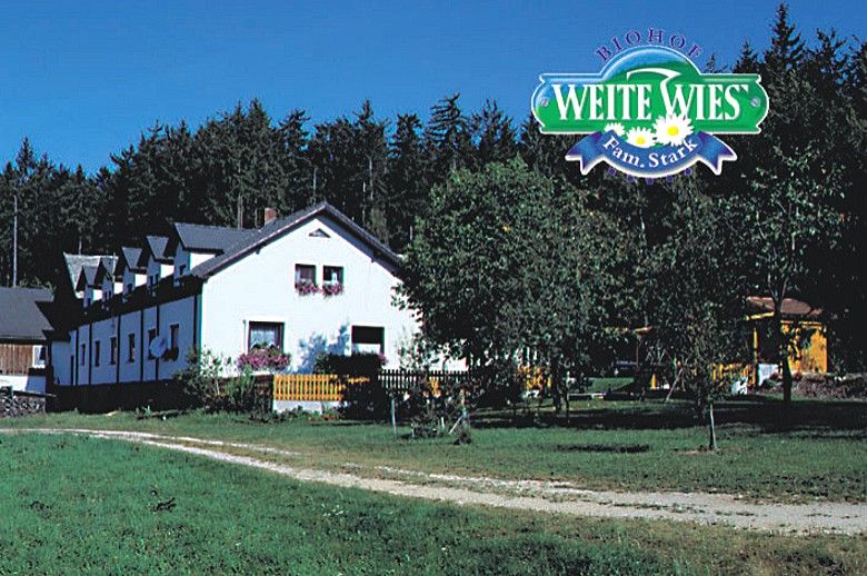 Bílý statek s květinami před lesem, nad nímž je logo "Biohof Weite Wies Fam. Stark".
