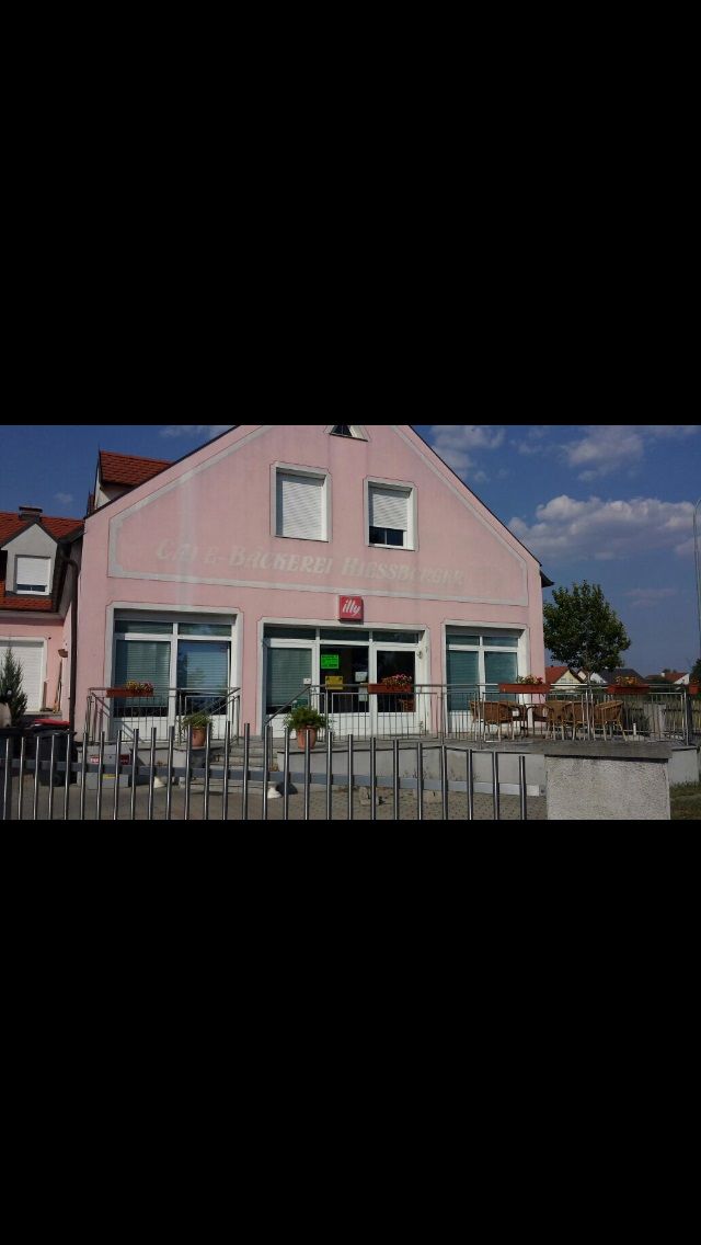 Pohled zvenčí na růžovou budovu s nápisem "Bäckerei-Café Hiessberger". Před budovou jsou stoly a židle.