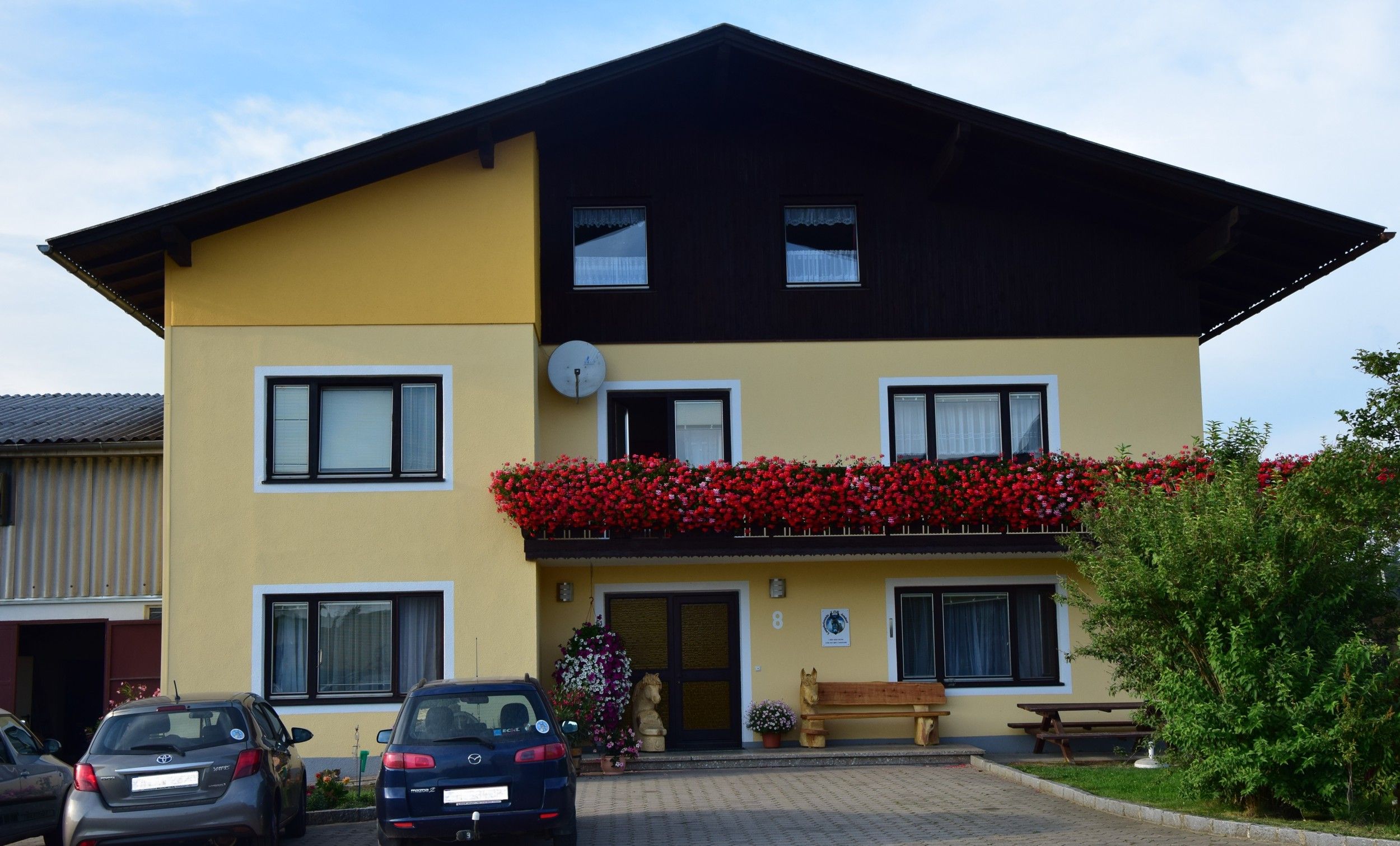 Pohled zvenčí na žlutý dům s balkonem plným červených květin a zaparkovanými auty před ním.