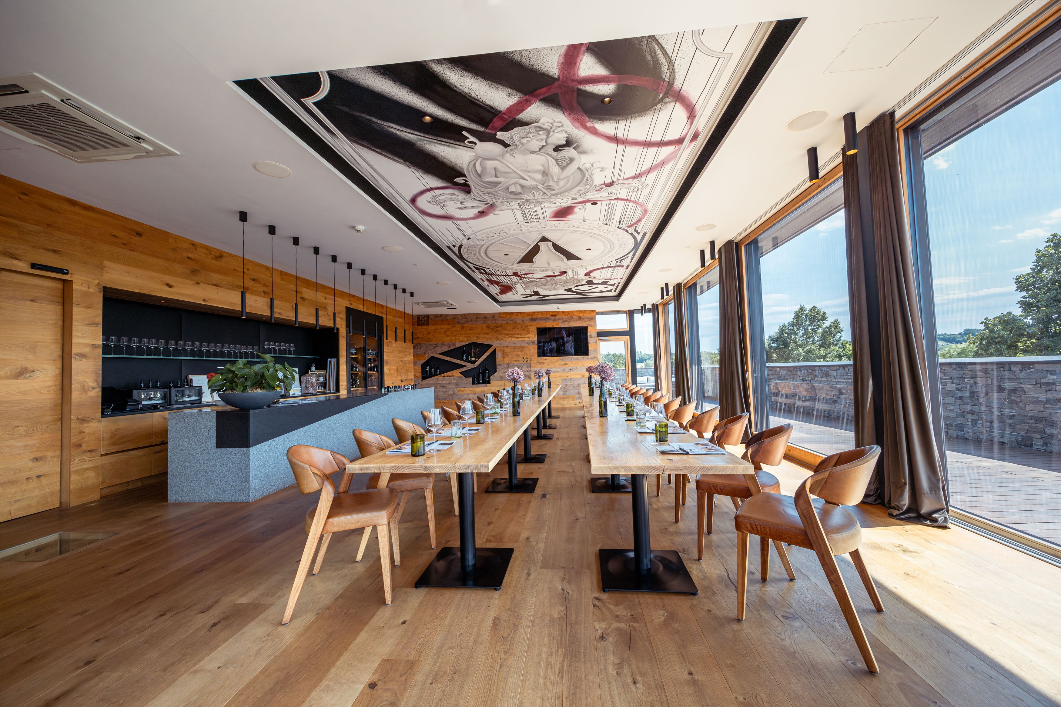 Pohled do interiéru moderní restaurace s dlouhými dřevěnými stoly, koženými křesly a velkými okny.