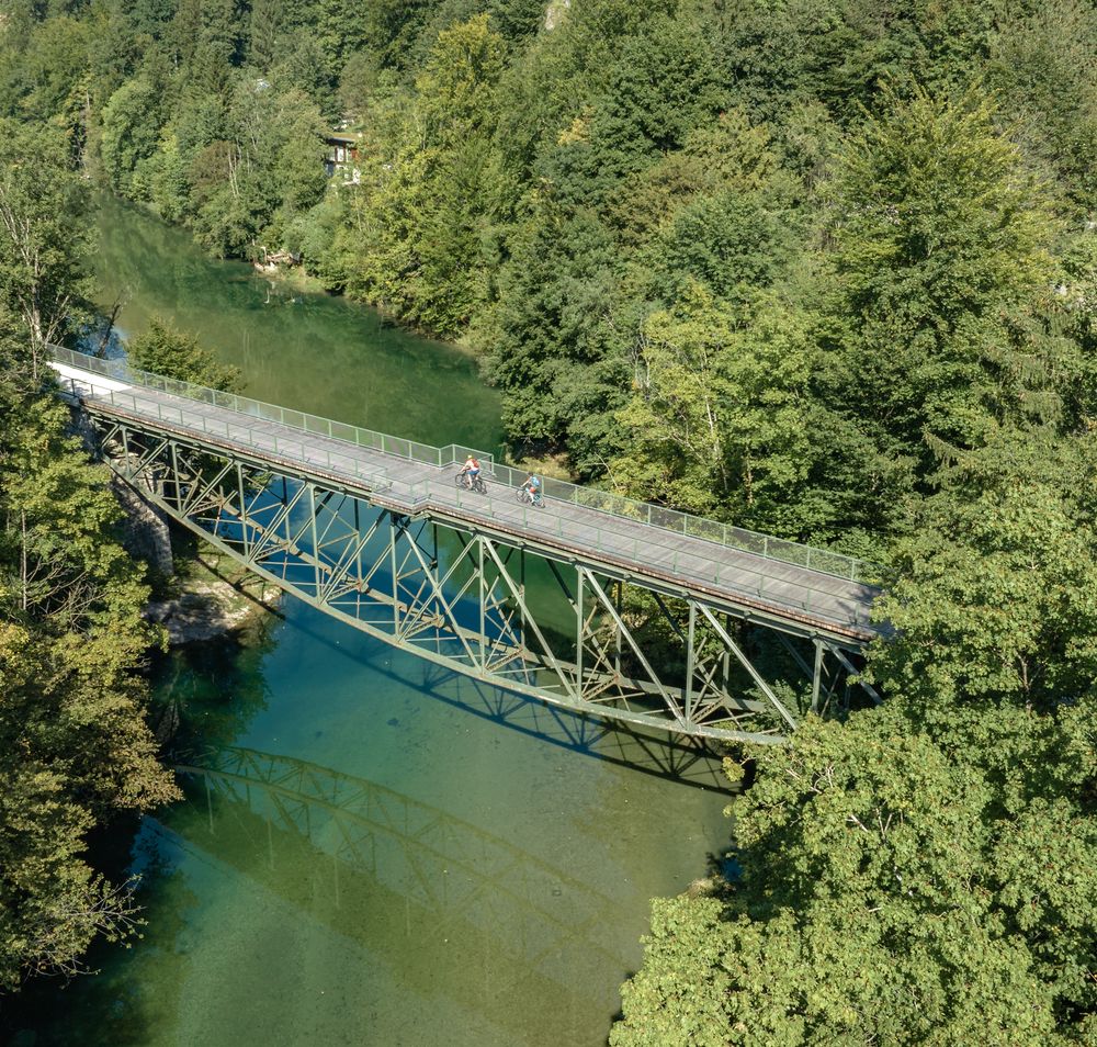 Eine malerische Brücke schwingt sich über das glitzernde Wasser, umgeben von üppigem Grün und majestätischen Bergen. Radfahrer genießen die frische Luft und die atemberaubende Aussicht auf die Ybbstaler Alpen, während sie die Ruhe der Natur erleben.
