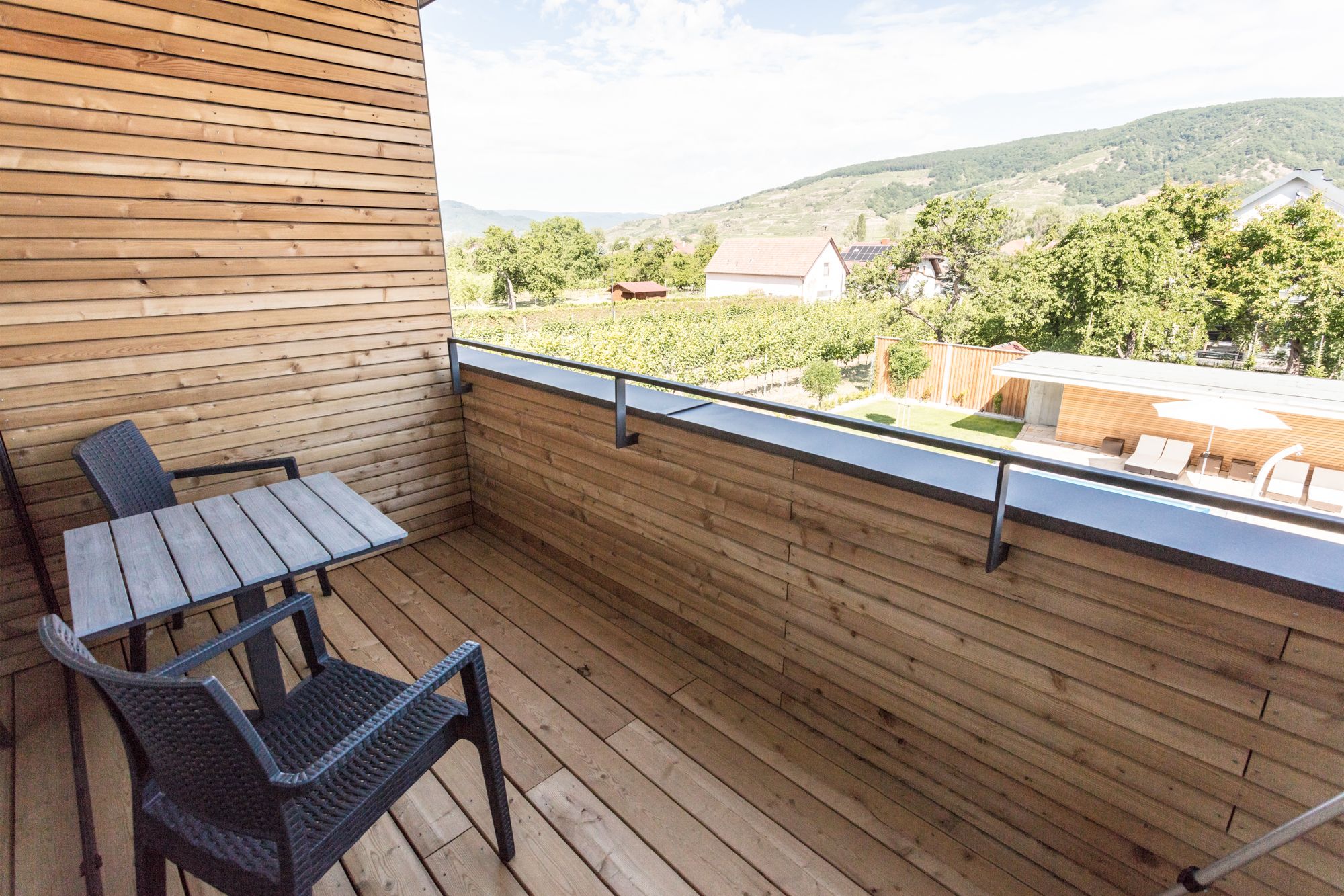 Dřevem obložený balkon se stolem a dvěma židlemi, výhled na zelenou krajinu a kopce v pozadí.