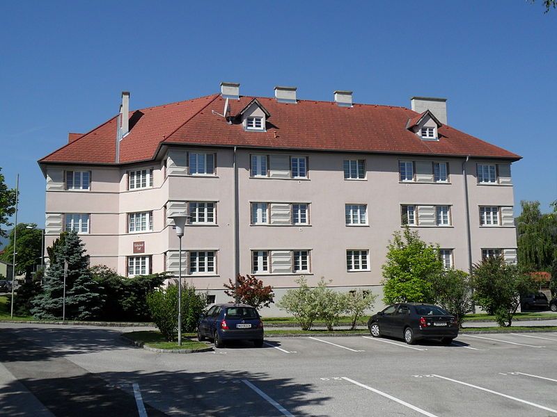 Obytná budova s červenou střechou a několika okny, obklopená stromy a zaparkovanými auty.