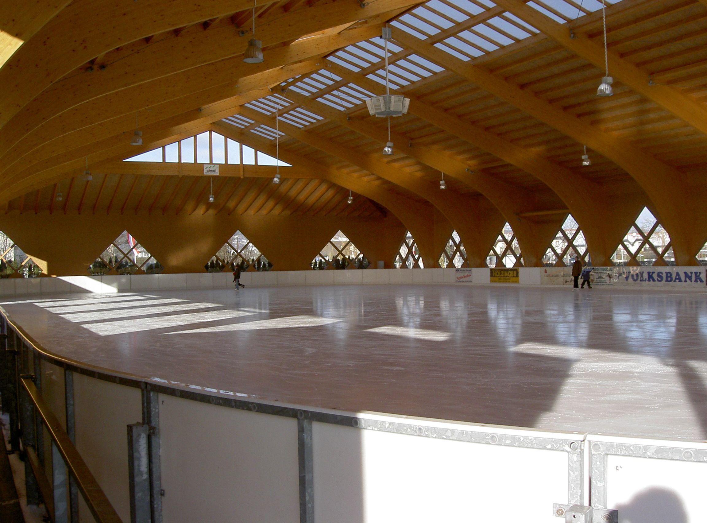 Pohled do interiéru krytého umělého kluziště s dřevěnou střechou a několika lidmi na ledě.