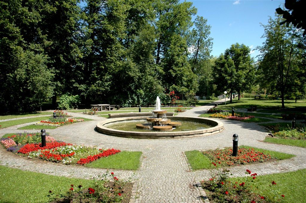 Udržovaný park s centrální fontánou, obklopený květinovými záhony a stromy.