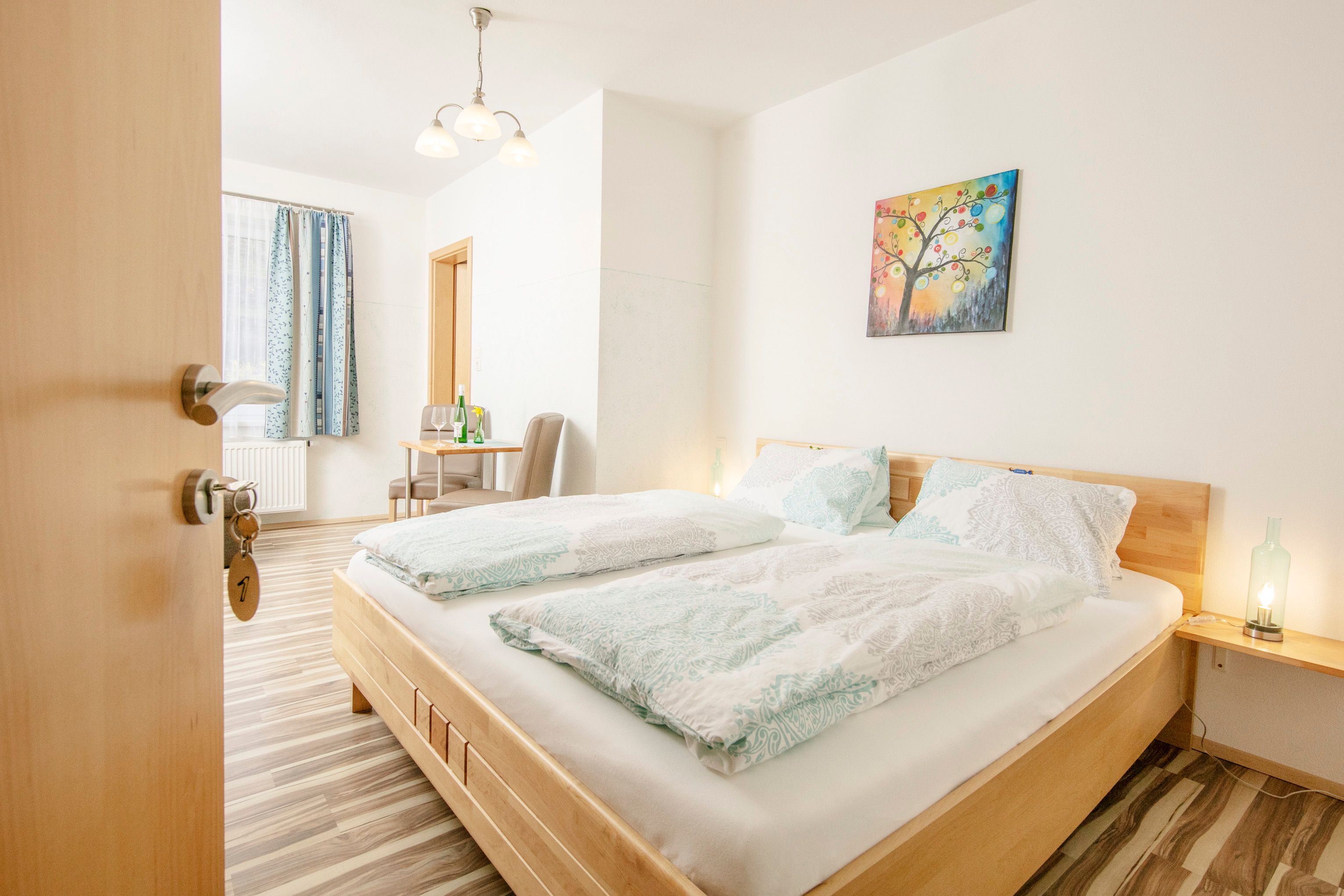 Světlý pokoj pro hosty s manželskou postelí, stolem, židlemi a barevnou nástěnnou malbou.