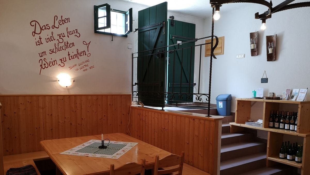 Pohled do interiéru útulného vinného sklípku s dřevěnými stoly a policemi, který zdobí Goethův citát na zdi.