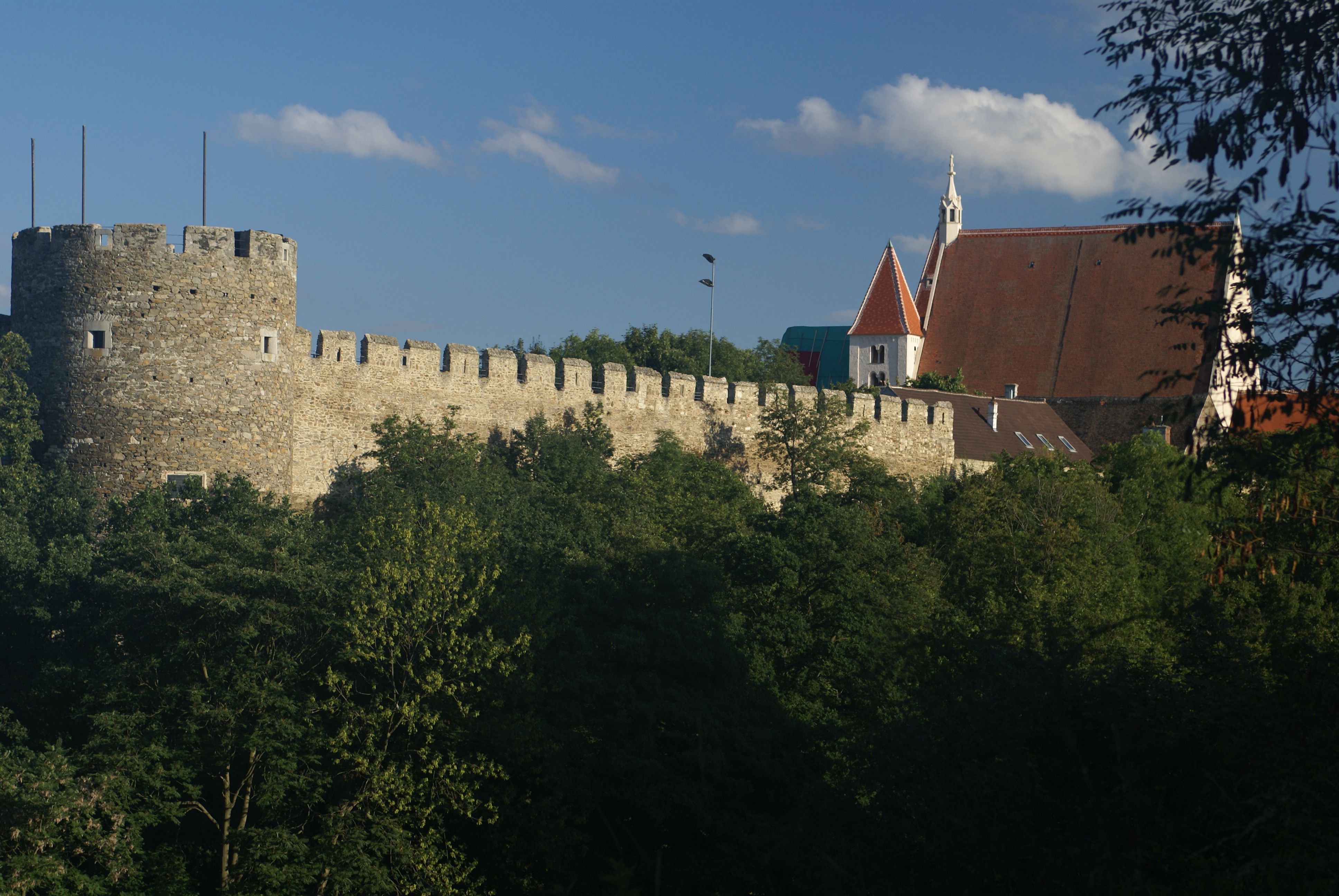Městské hradby s kancléřskou věží a farním kostelem v pozadí, obklopené stromy.