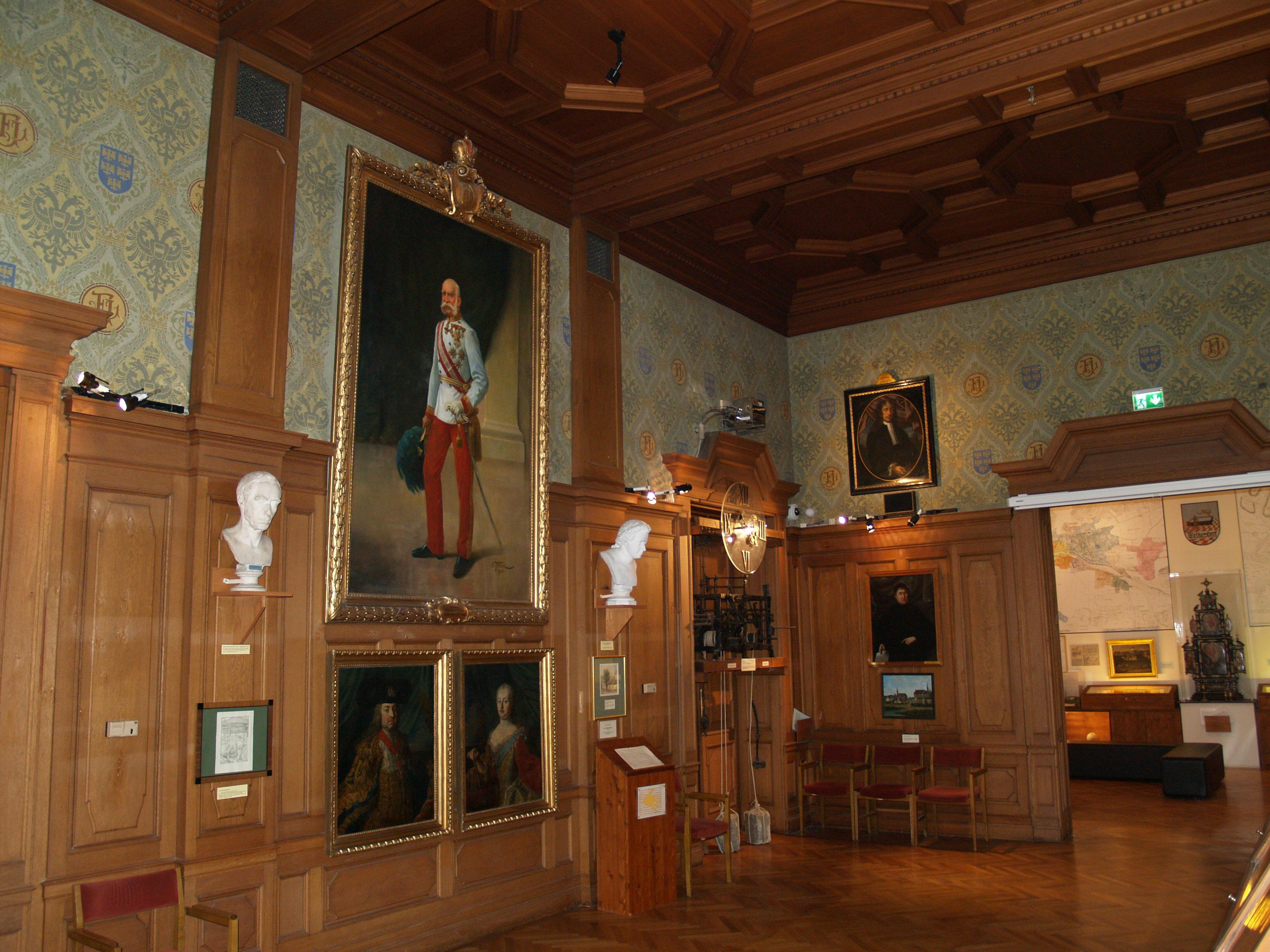 Historický sál s dřevěným obložením, obrazy a bustami na stěnách.