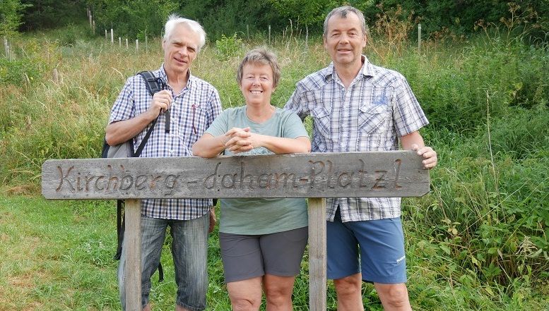 Tři lidé stojí za dřevěnou cedulí s nápisem "Kirchberg-daham-Platzl".