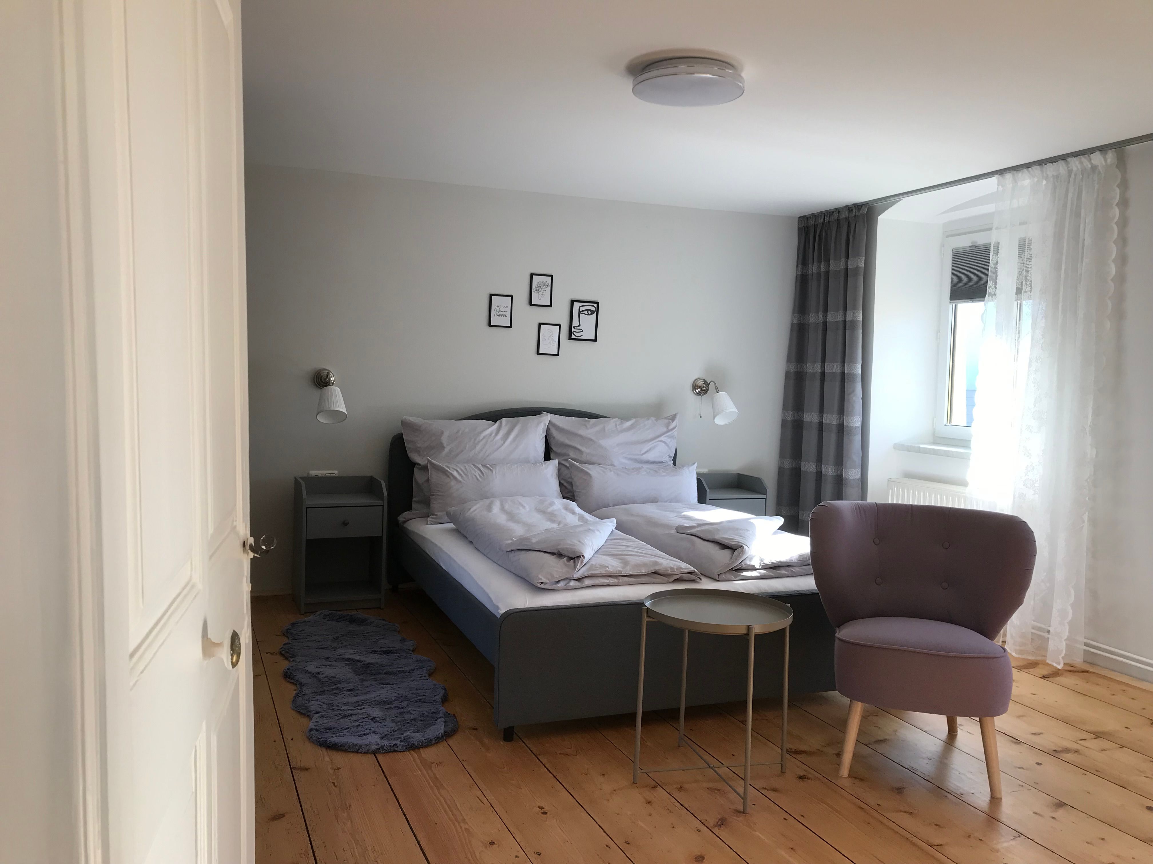 Moderně zařízený dvoulůžkový pokoj s postelí, křeslem a malým stolkem na dřevěné podlaze.