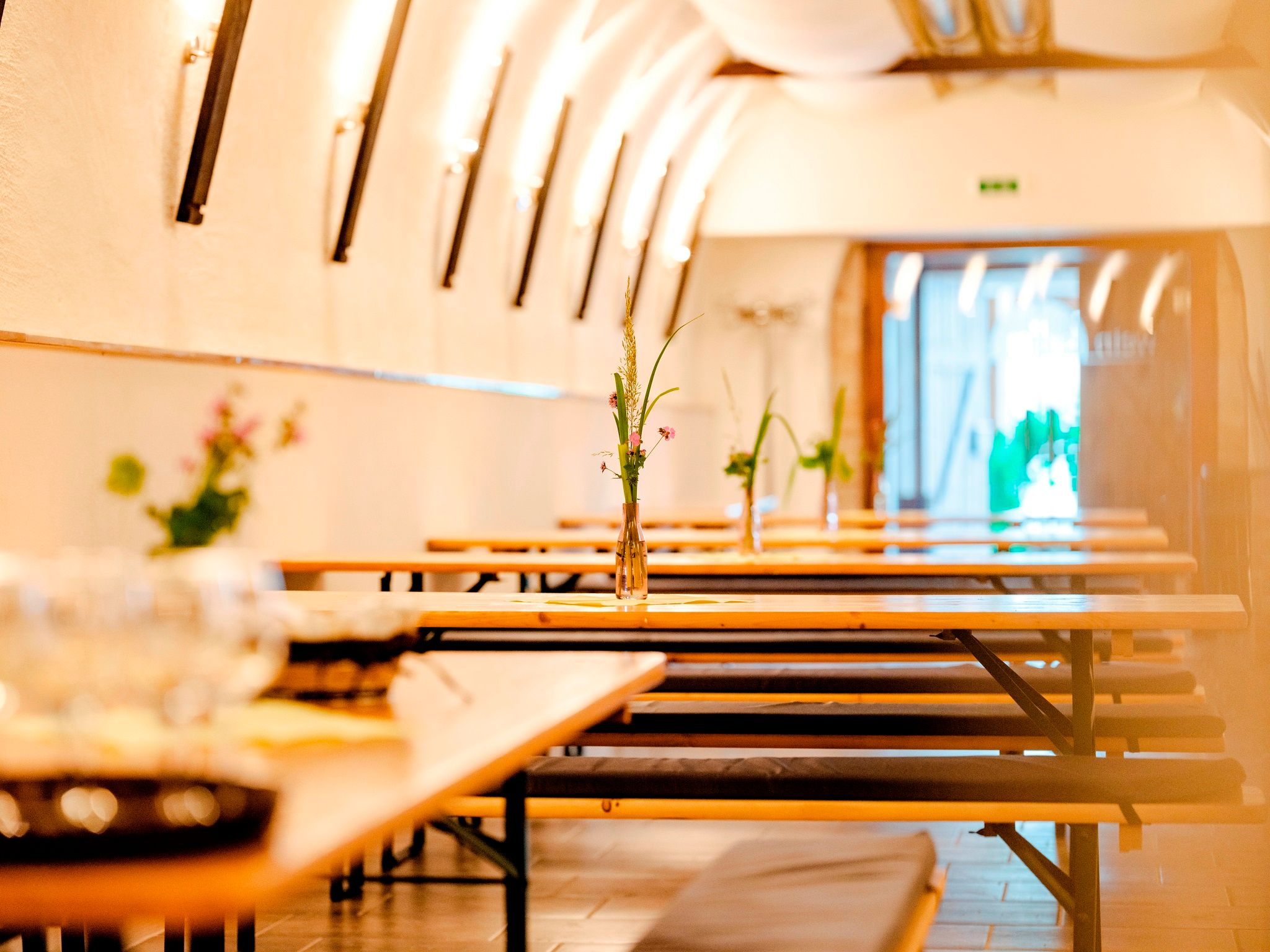 Pohled do interiéru vinného šenku s dlouhými dřevěnými stoly a lavicemi, zdobenými vázami s květinami.