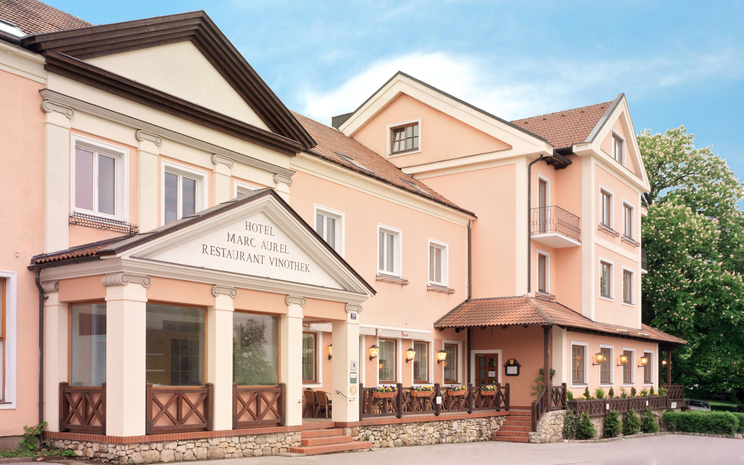 Pohled zvenčí na hotel Marc Aurel v Petronell-Carnuntum s růžovou fasádou a klasickým vchodem.