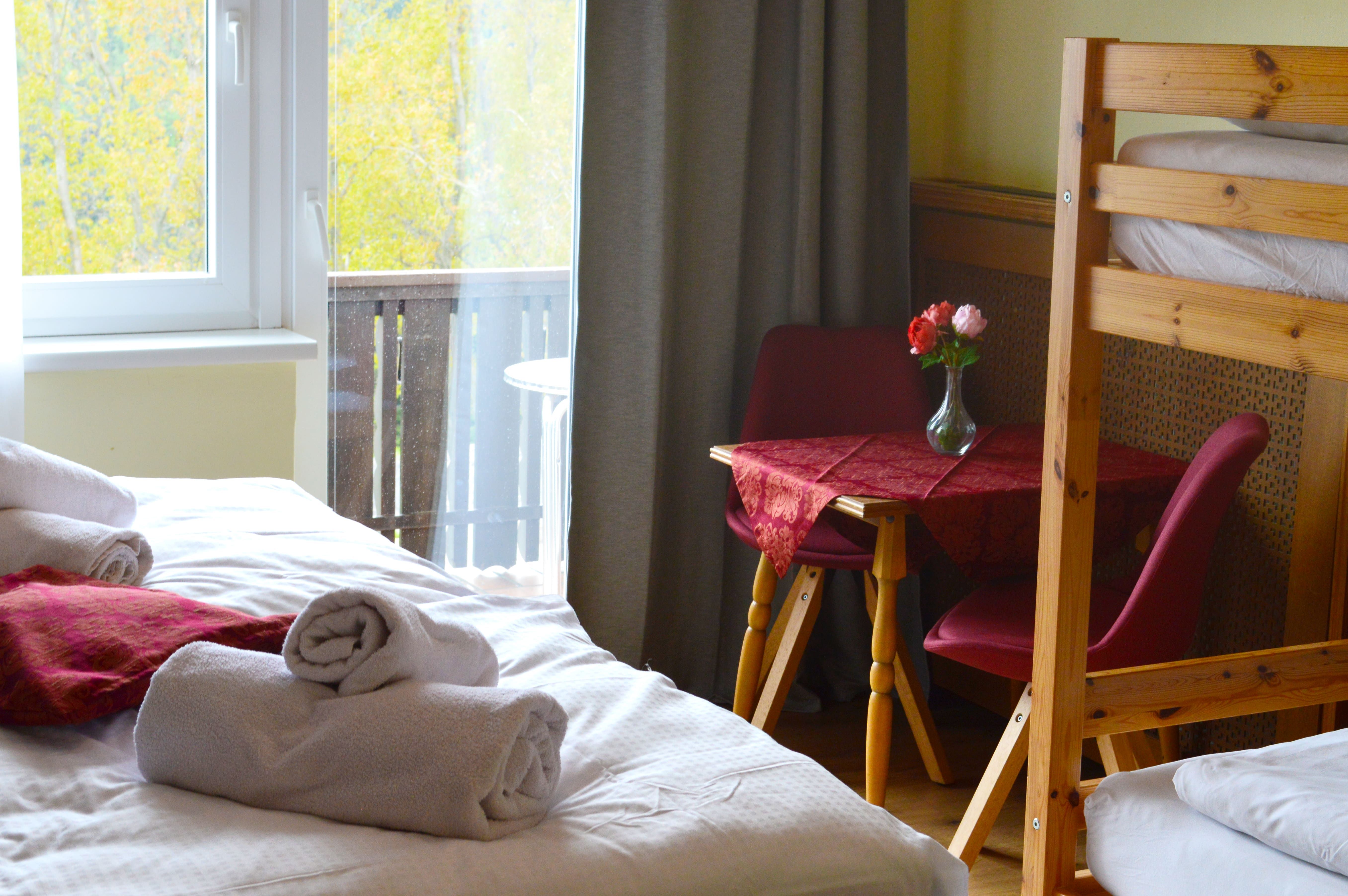 Útulný pokoj s postelí, stolem s červenými židlemi a kyticí květin. Z okna je vidět balkon a podzimní stromy.