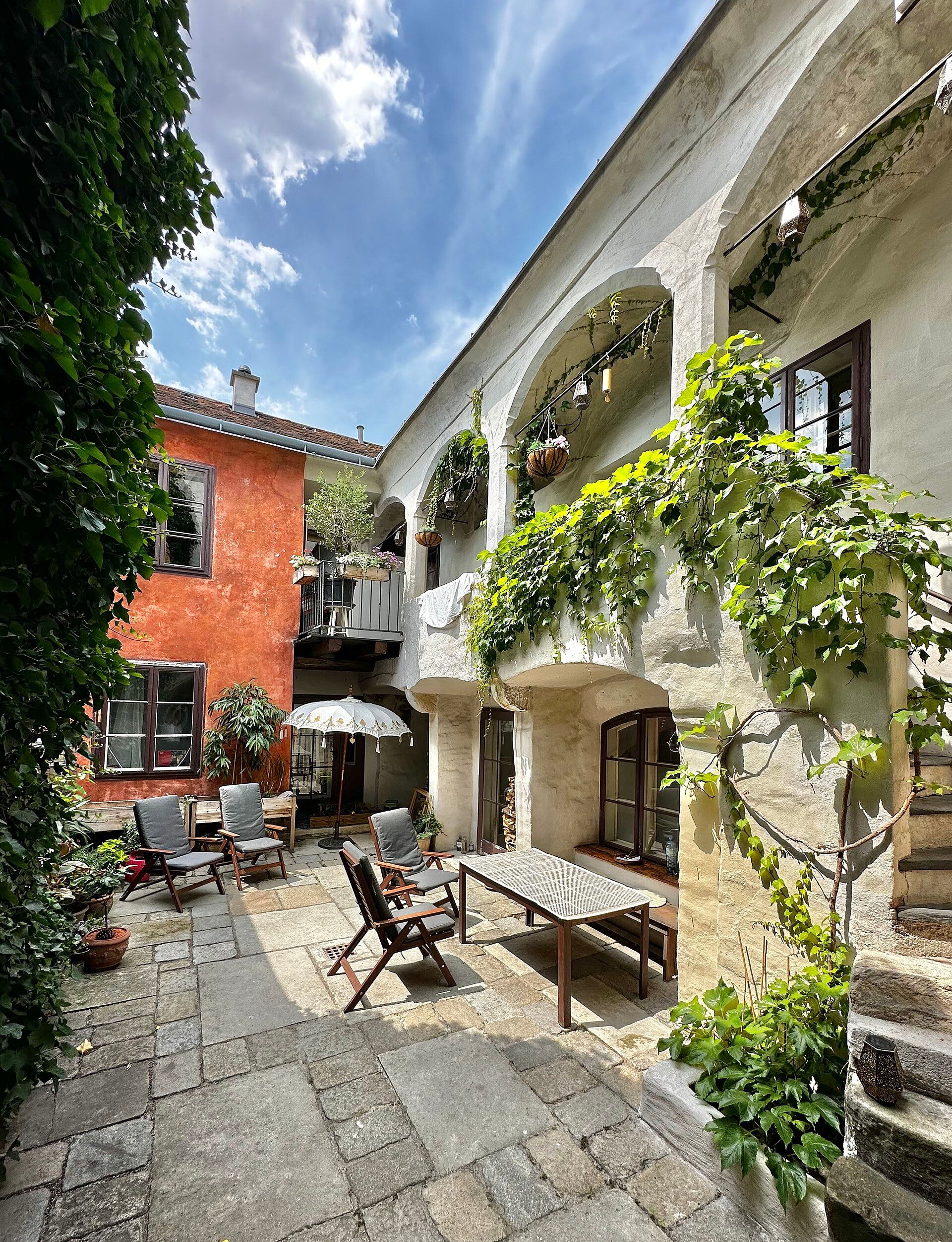 Útulný dvůr s rostlinami, dřevěným nábytkem a slunečníkem.