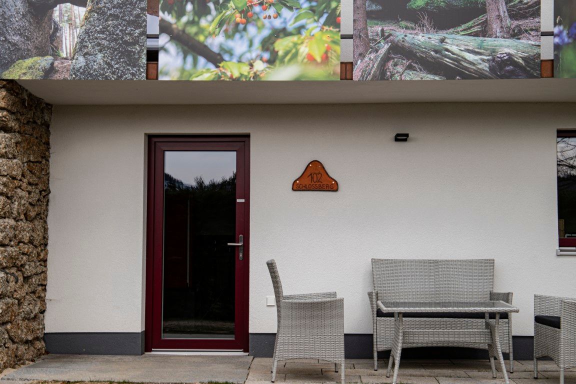 Vstupní část budovy s dveřmi, cedulí s nápisem „102 Schlossberg“ a zahradním nábytkem před ní.