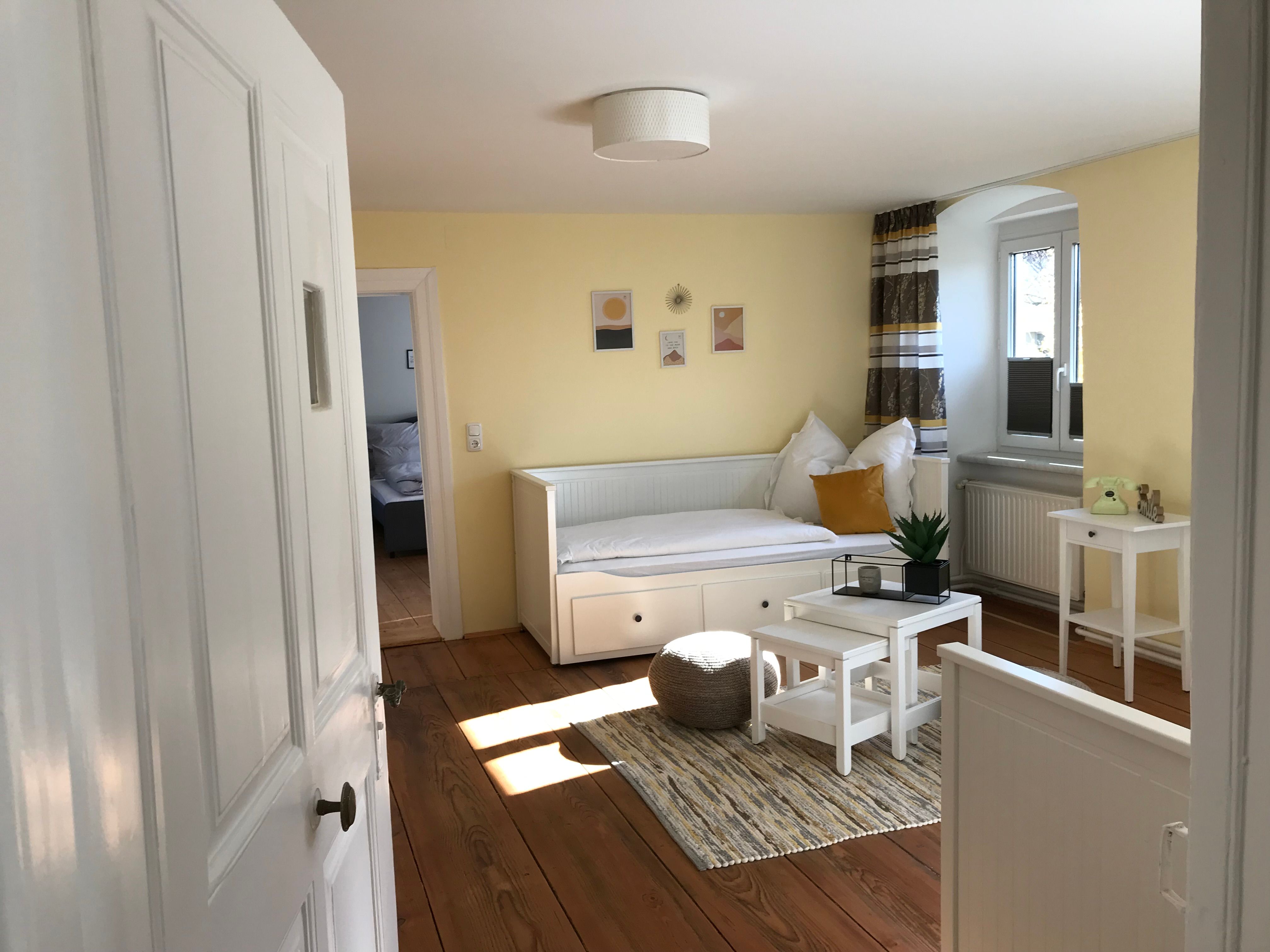 Světlý pokoj se žlutými stěnami, bílou denní postelí, malým stolkem a rostlinami.