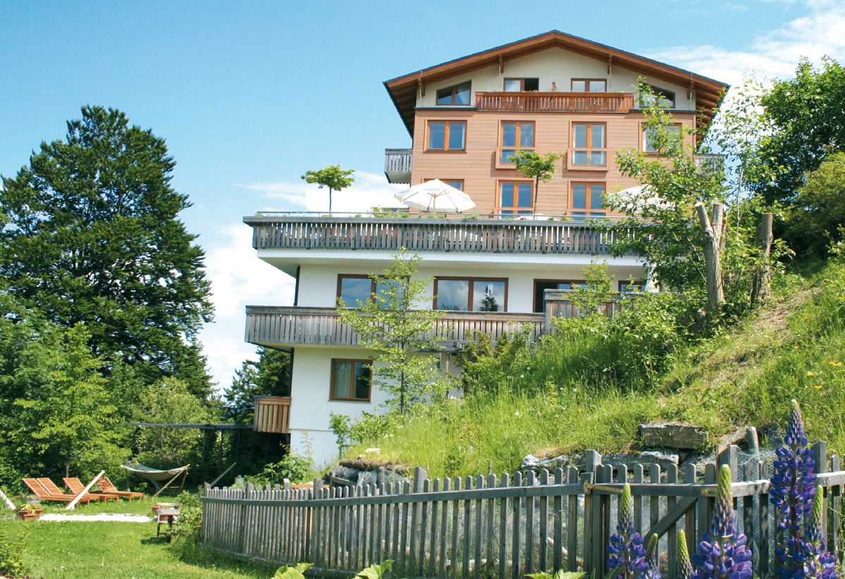 Vícepodlažní hotel s dřevěnými balkony, obklopený zelenou přírodou a kvetoucími květinami, pod modrou oblohou.