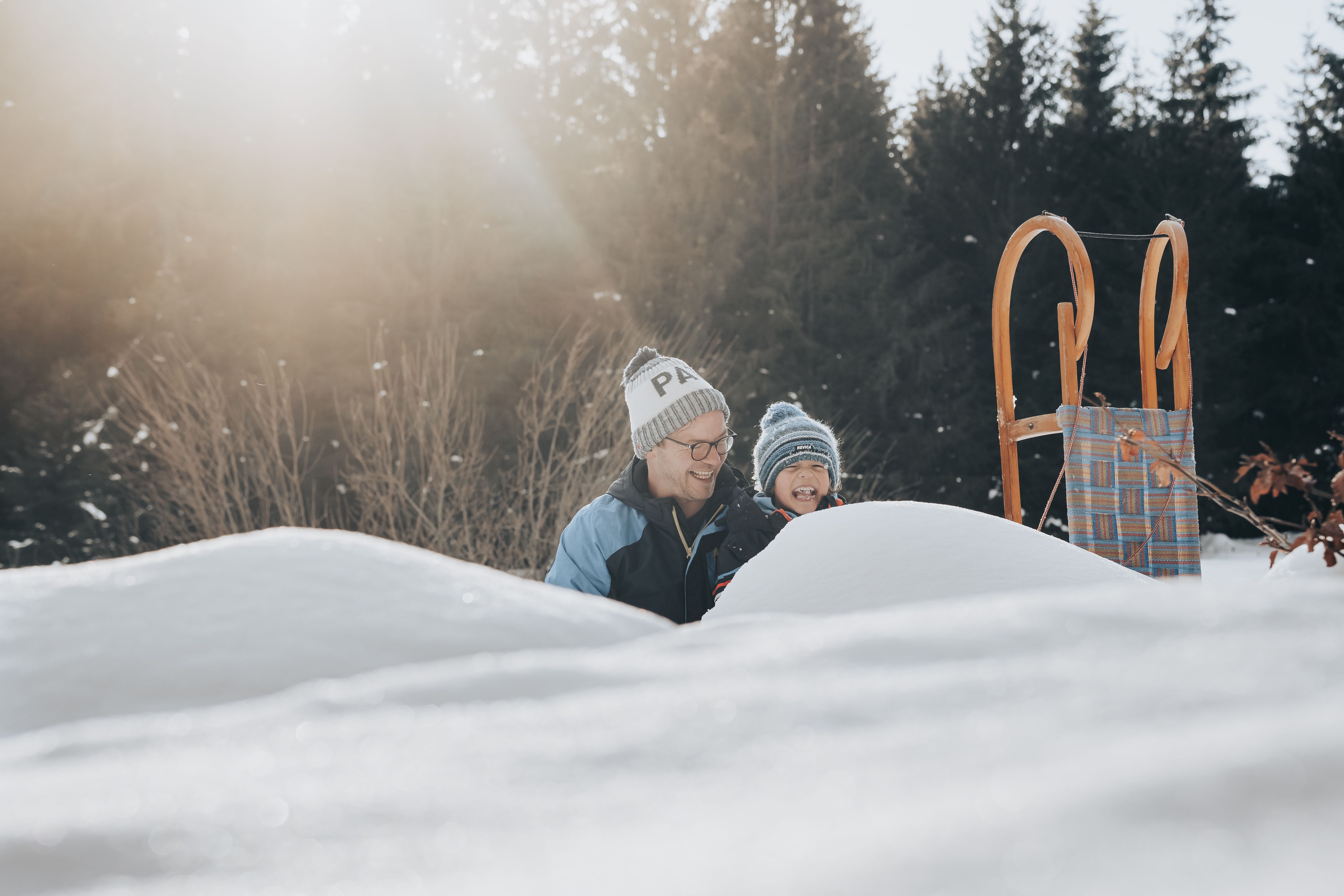 Eine fröhliche Familie genießt den Winter in der verschneiten Landschaft der Ybbstaler Alpen. Lachen und Freude erfüllen die Luft, während sie gemeinsam mit einem Schlitten die sanften Hügel hinunterrodeln. Die strahlende Sonne und der glitzernde Schnee schaffen eine magische Atmosphäre.