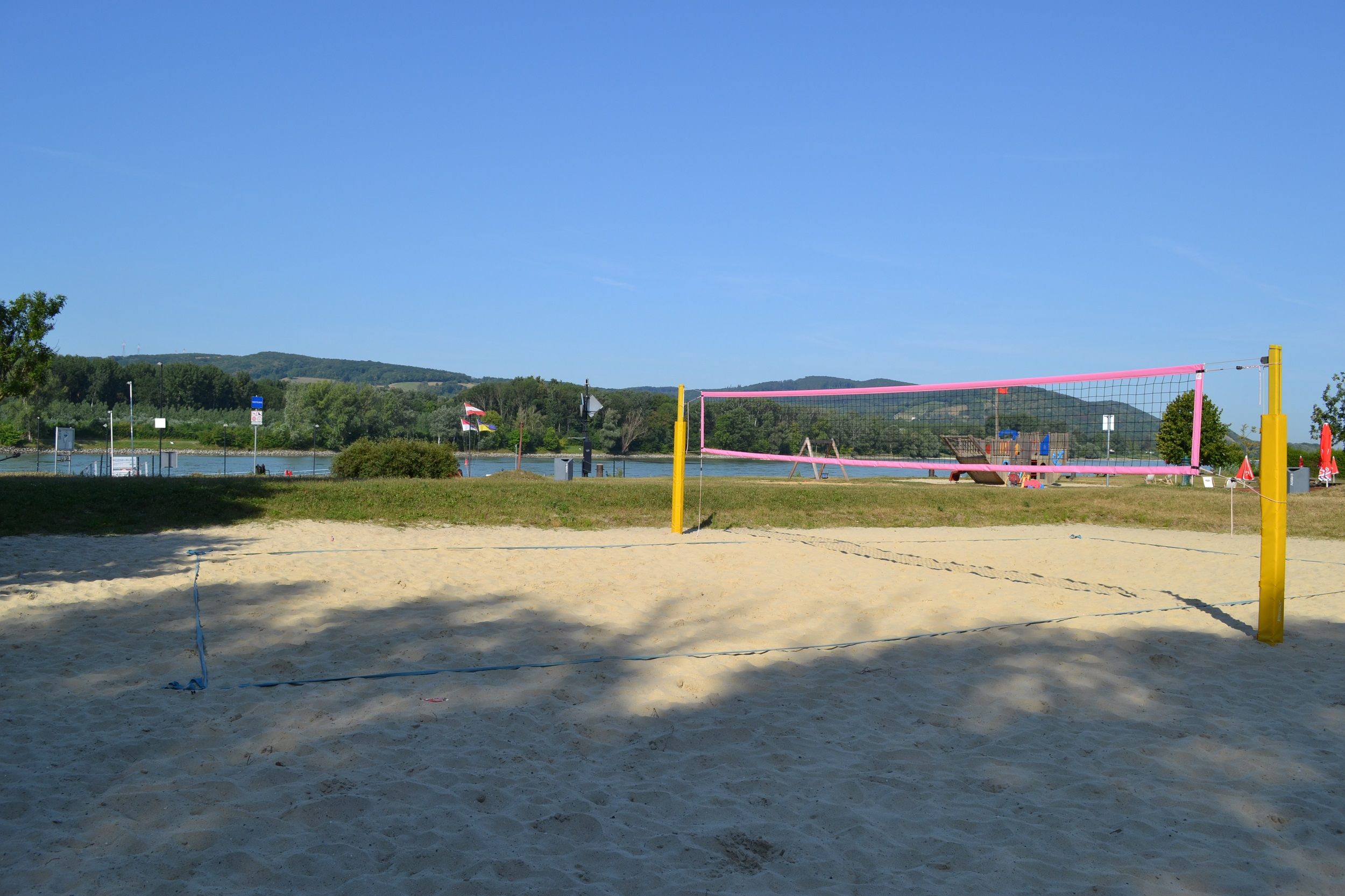 Prázdné hřiště na plážový volejbal se žlutými sloupky a růžovou sítí před řekou a zelenými kopci v pozadí.