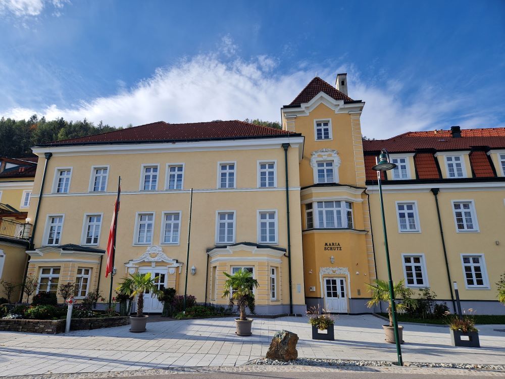 Žlutá budova s červenými střechami a několika okny, obklopená rostlinami a vlajkami.