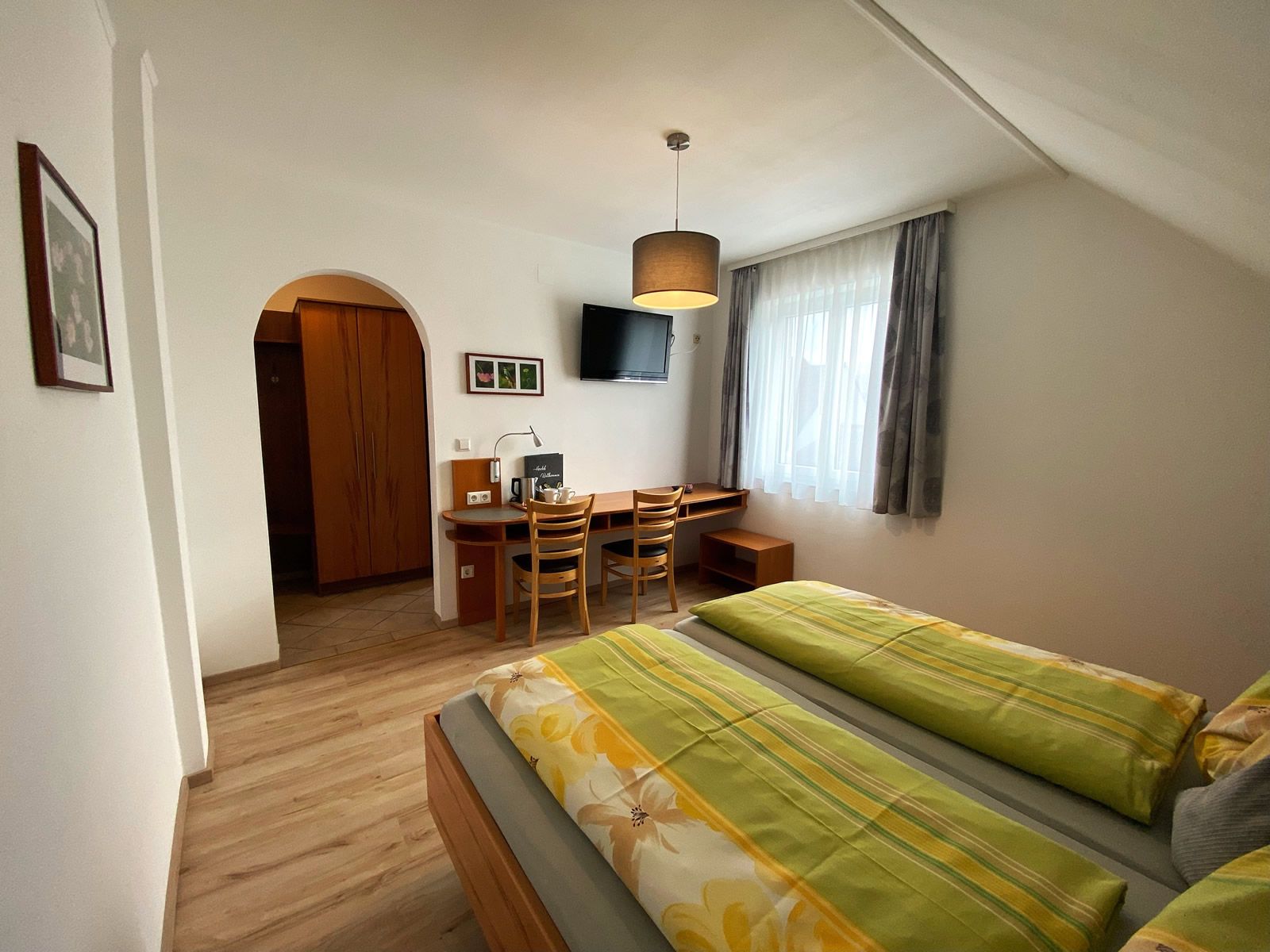 Útulný dvoulůžkový pokoj se dvěma lůžky, psacím stolem, židlemi a televizí na stěně.