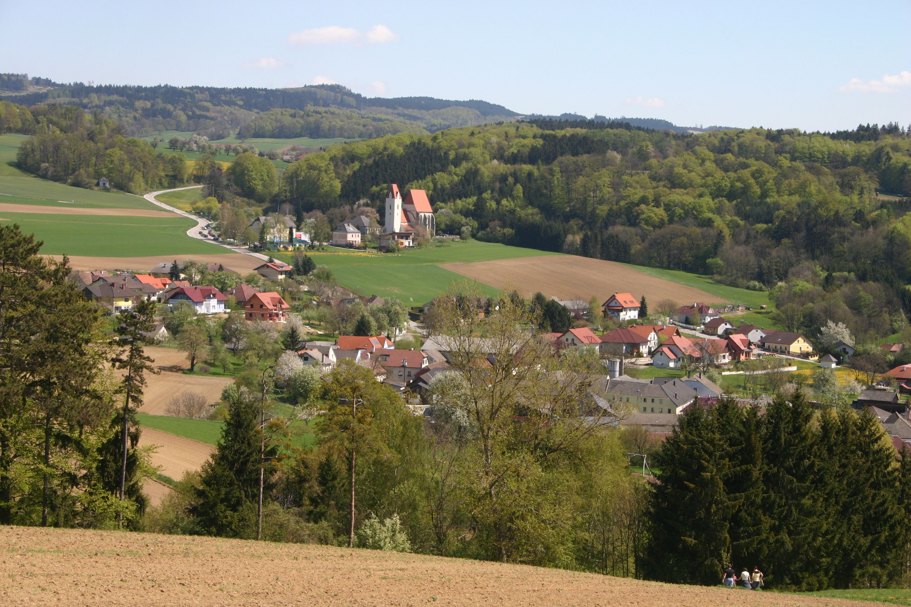 Krajina s vesnicí, kostelem a kopci v pozadí.