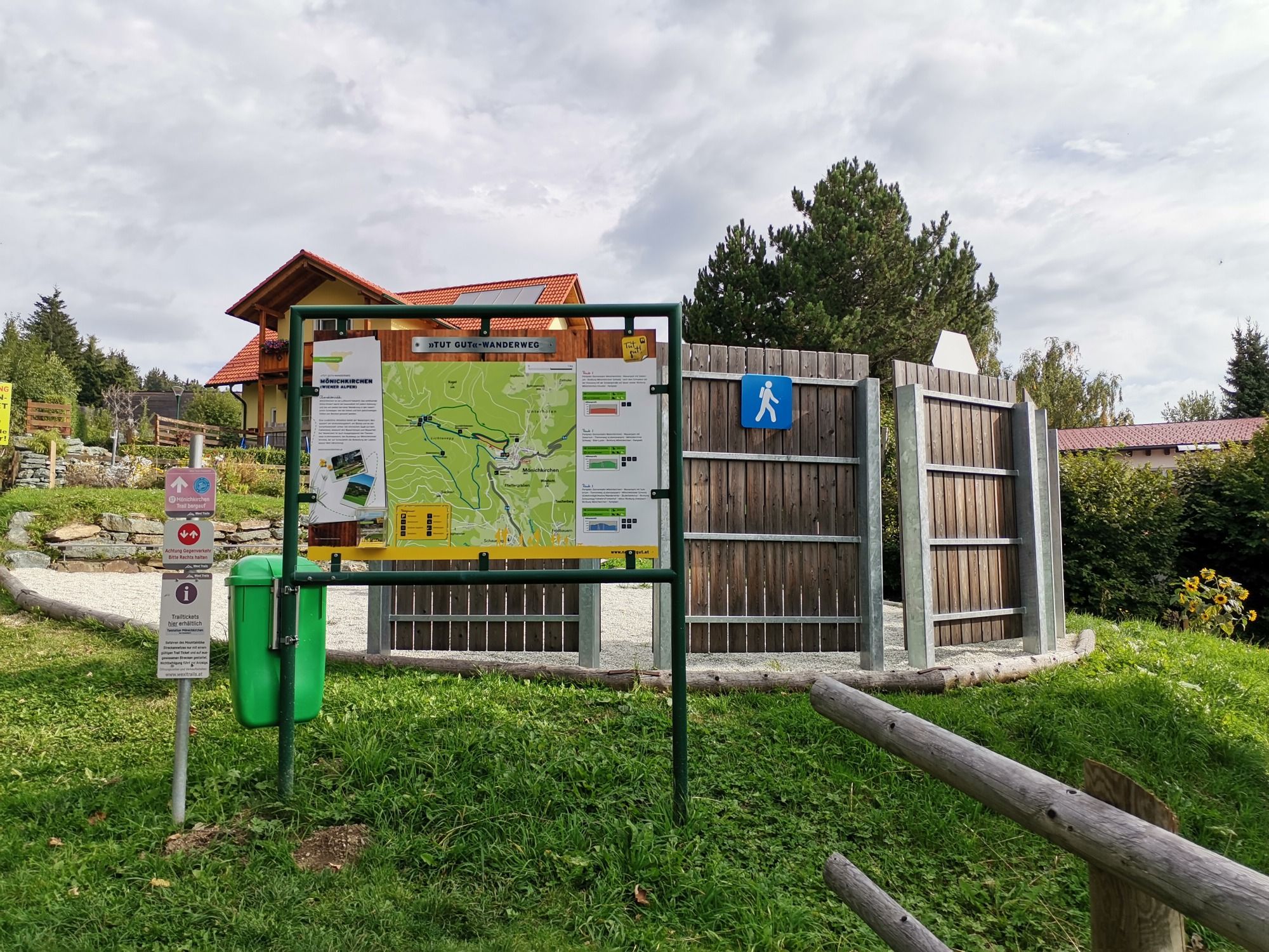 Informační tabule turistické stezky s mapou a značkami na louce, obklopené stromy a budovou v pozadí.