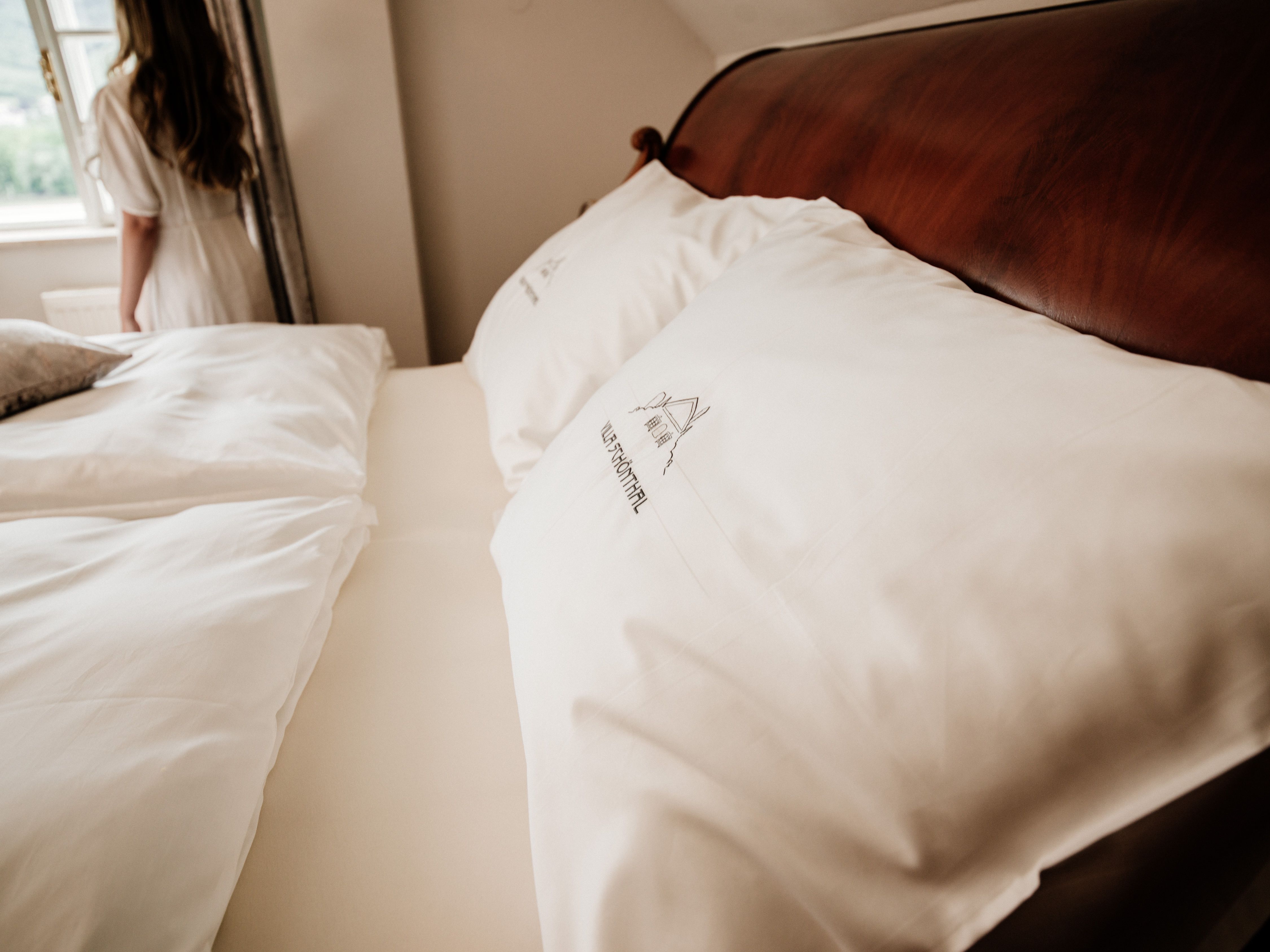 Detailní záběr na postel s bílými polštáři a ložním prádlem, v pozadí žena u okna.