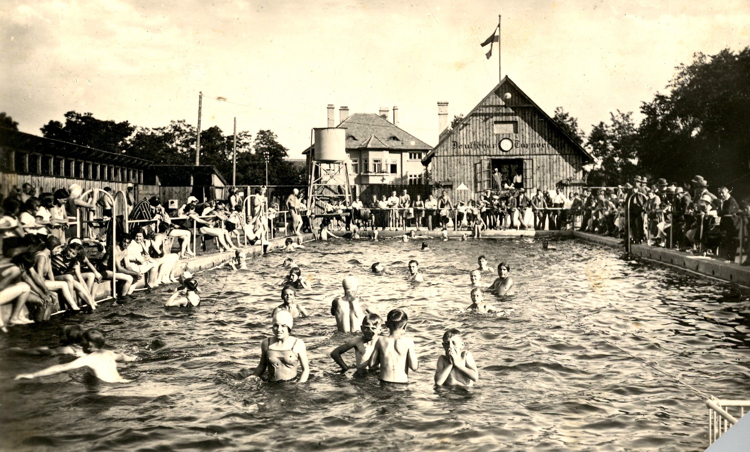 Historická fotografie venkovního koupaliště s mnoha lidmi ve vodě i u okraje bazénu. V pozadí je budova s vlajkou.