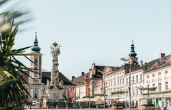 Die sonnendurchflutete Platzlandschaft lädt zum Verweilen ein, während die historischen Gebäude im Hintergrund eine charmante Kulisse bieten. Palmen und bunte Blumen verleihen dem Ort eine lebendige Atmosphäre, perfekt für entspannte Sommertage.
