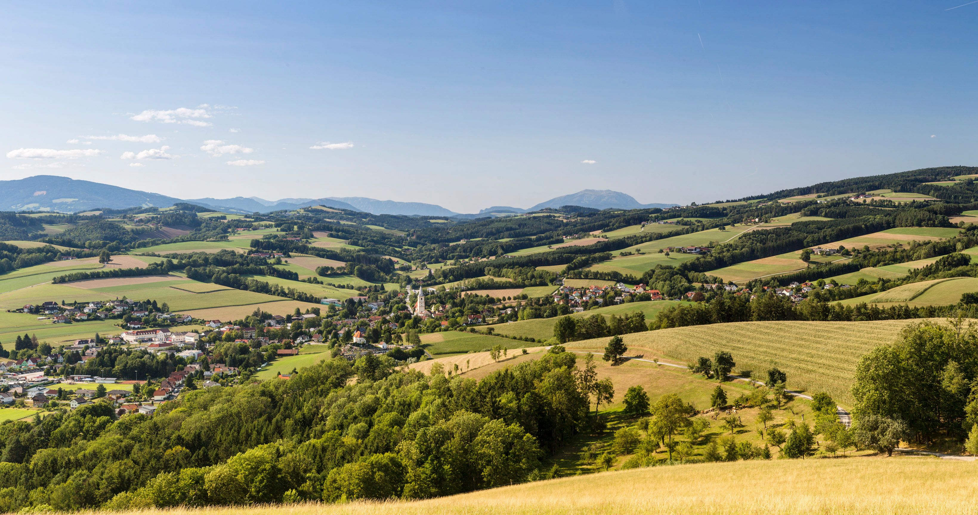 Panoramatický snímek z oblasti Buckelige Welt, v údolí leží městečko Krumbach s kostelní věží, v pozadí jsou kopce a hora Schneeberg.