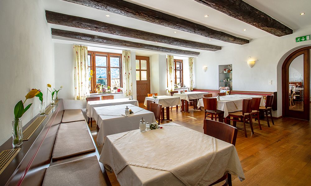 Útulná snídaňová místnost s dřevěnými trámy, prostřenými stoly a květinami na okenních parapetech.